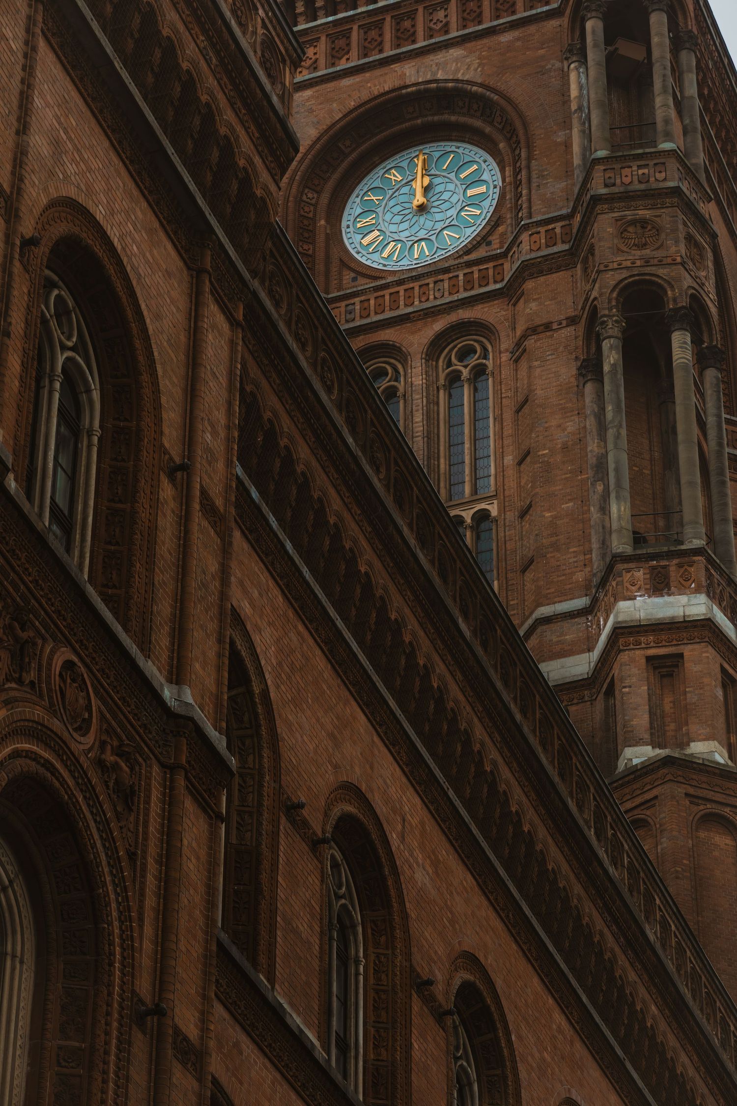 Historic brick building facade with ornate arches and illuminated blue clock face against warm brown stonework.