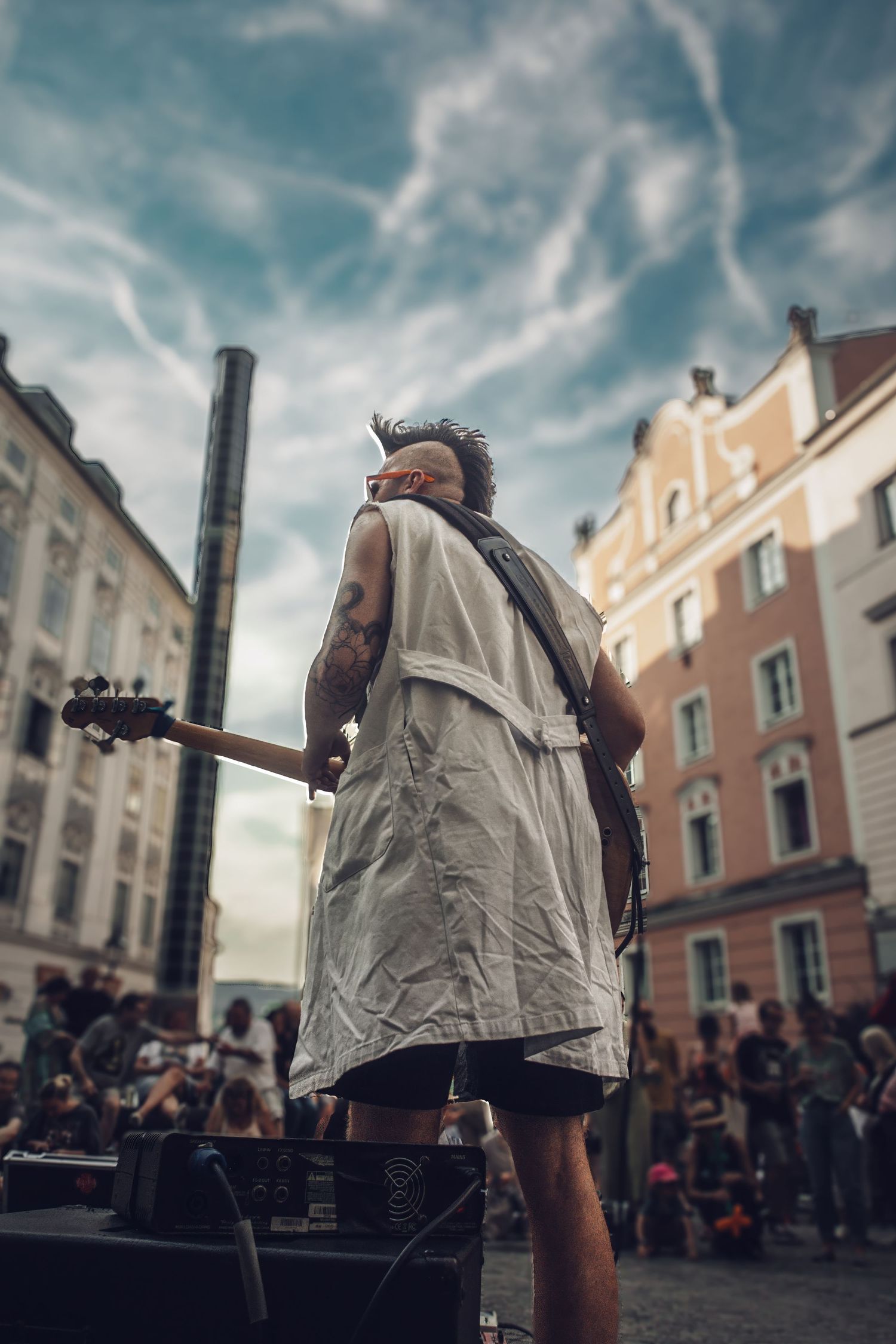 Performer on stage wearing white vest against urban backdrop with crowd below.