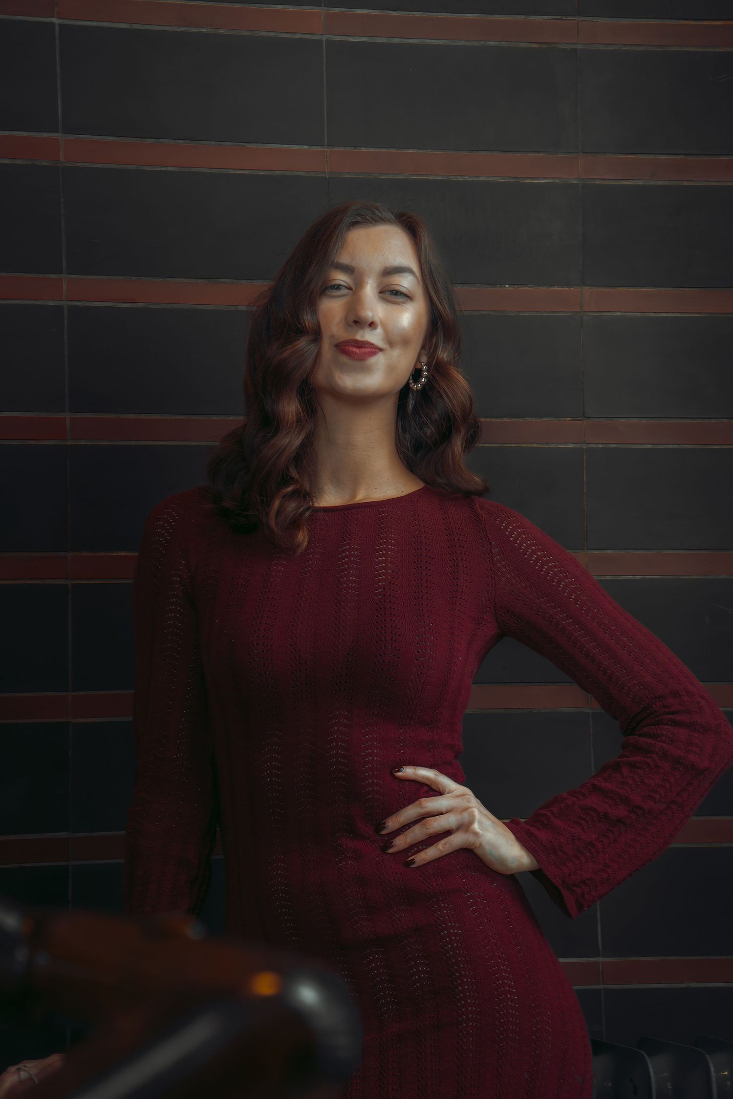Woman in burgundy knit dress poses elegantly against dark striped wall in moody studio lighting.