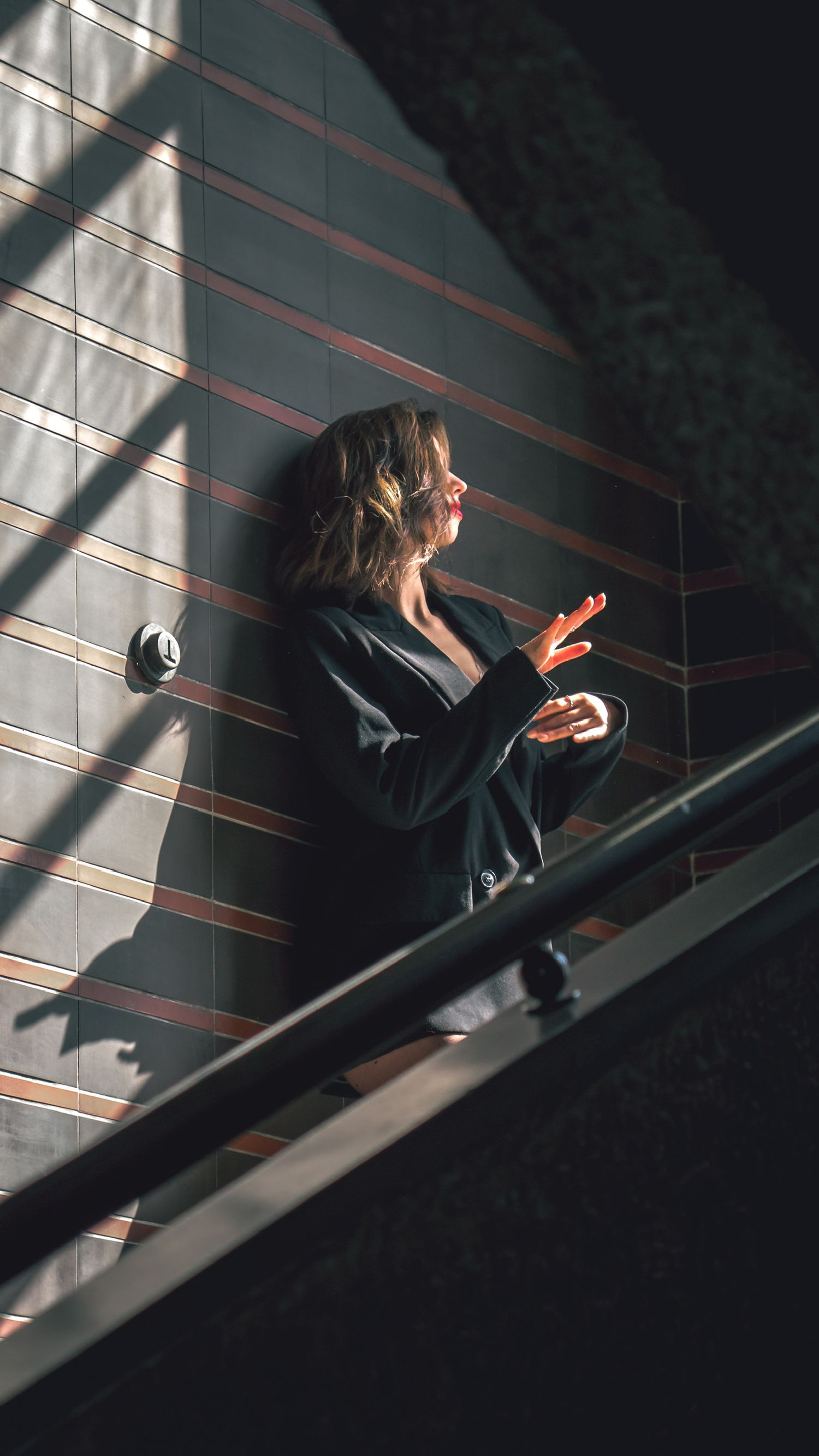 A person in dark clothing stands in a stairwell with dramatic shadows and light streaming through window blinds.