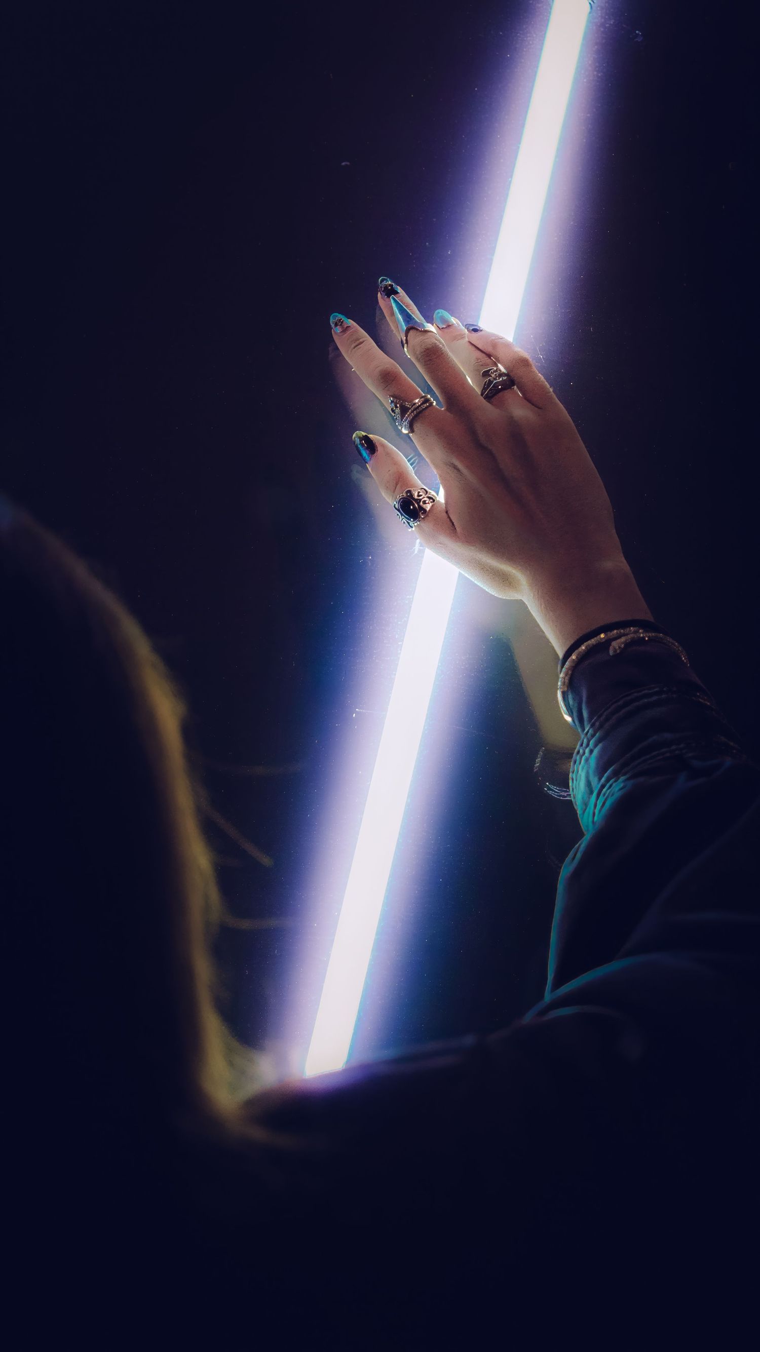 Hand with painted nails and rings reaching toward a bright vertical light beam against dark background.