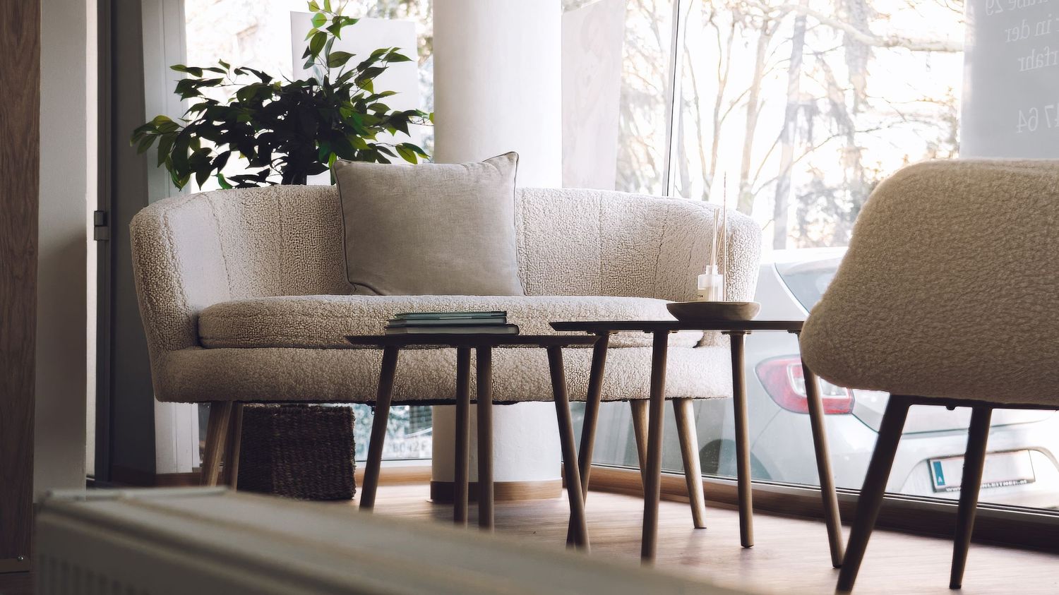 Modern living room interior with beige curved sofa, wooden side tables and a potted plant by the window.