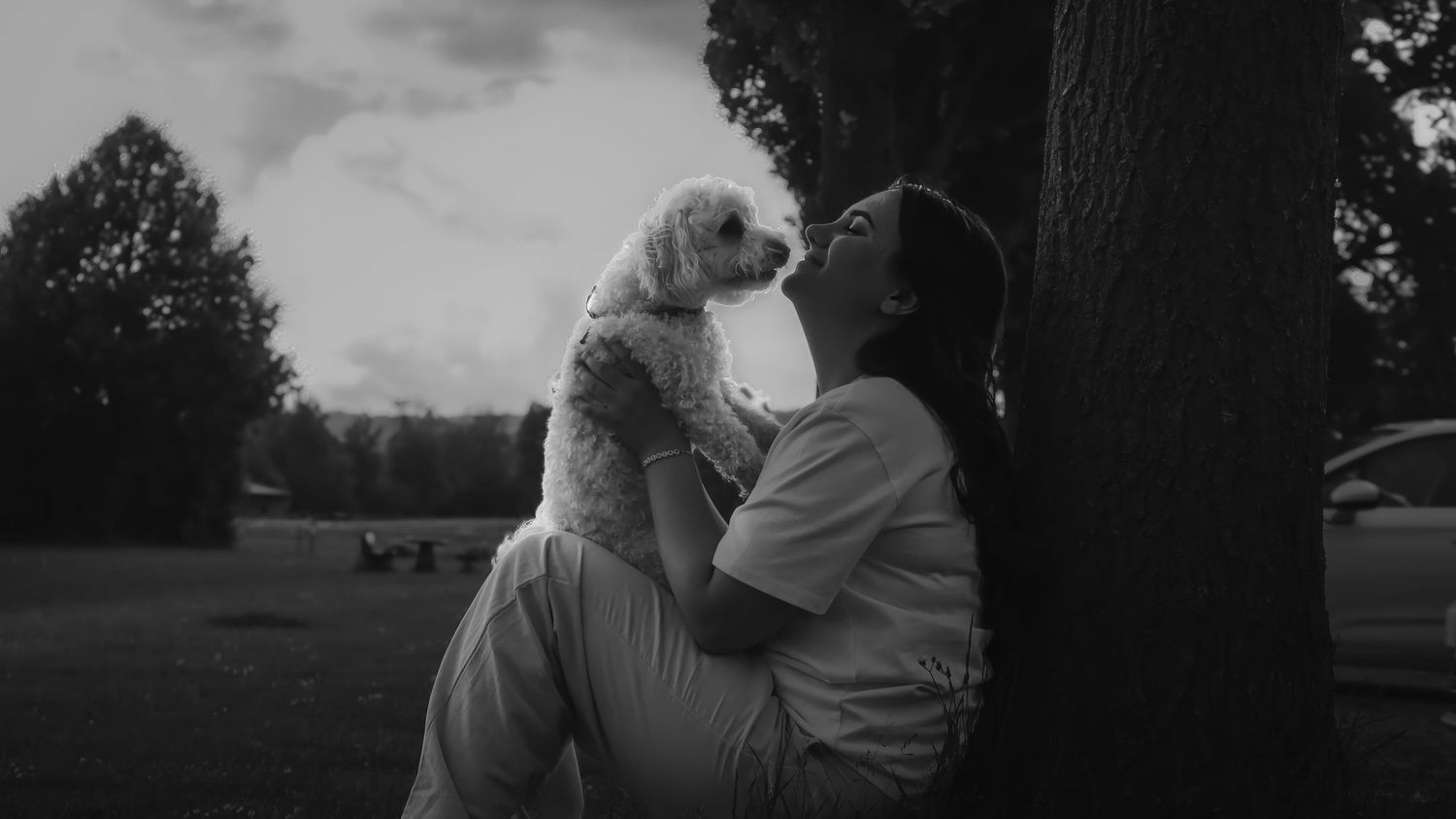 Black and white photo of a person holding a small dog while sitting under a tree in a park.