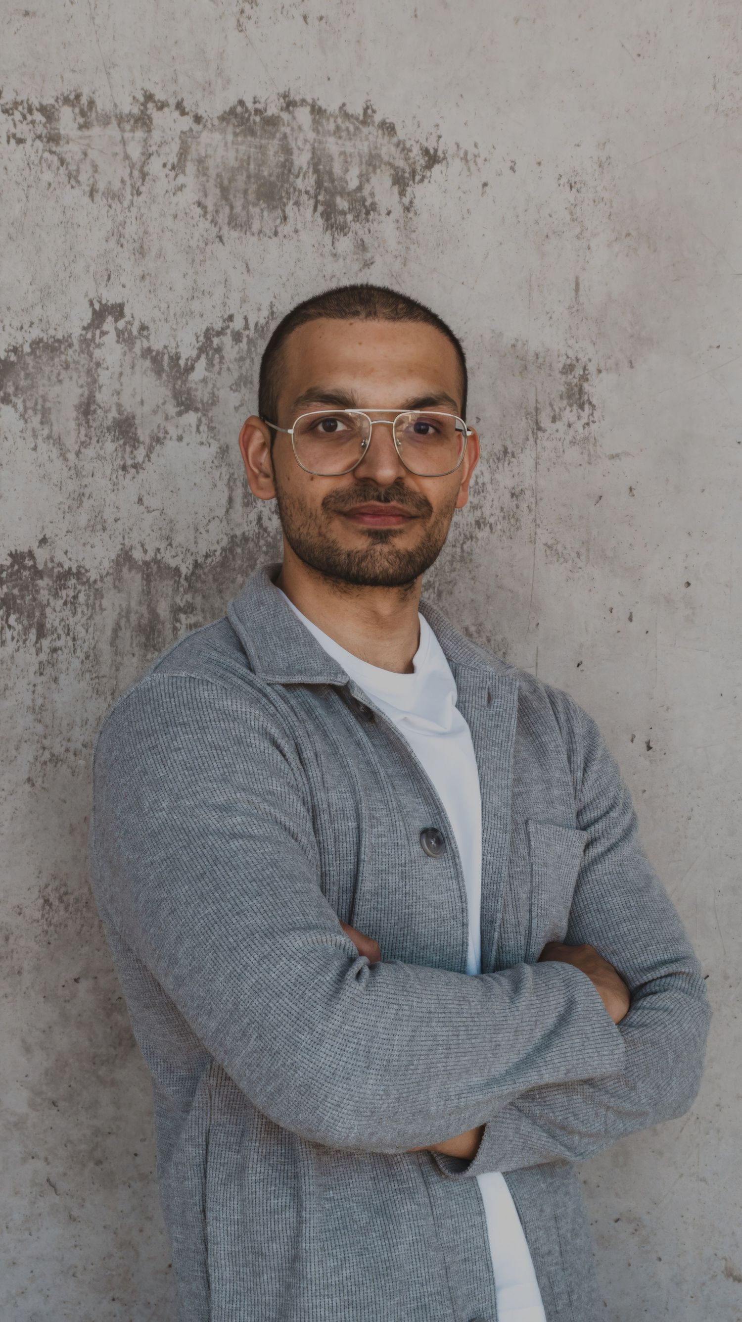 Professional portrait of someone wearing glasses and a grey cardigan sweater against a textured concrete wall.