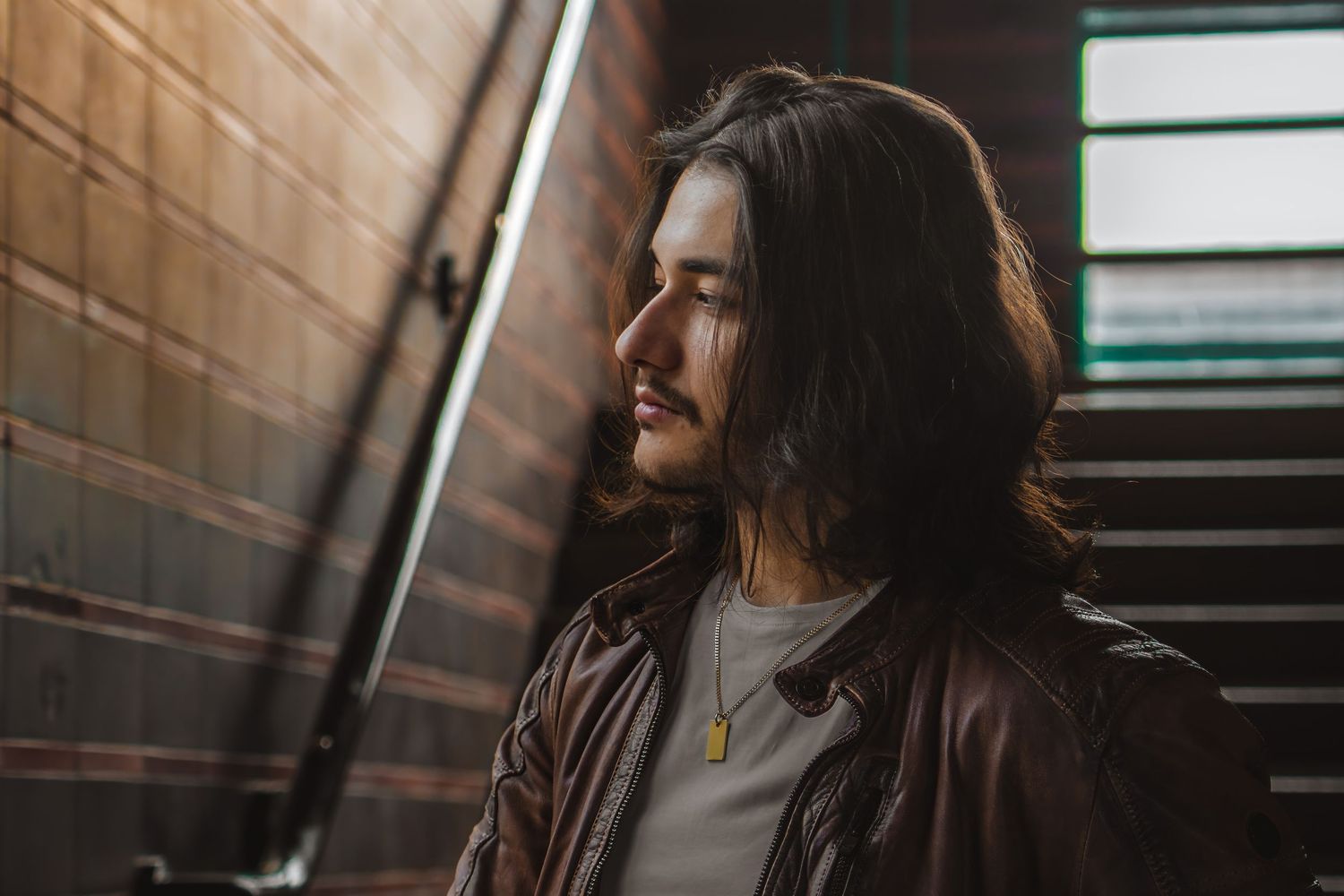 Person in leather jacket standing in stairwell with sunlit industrial backdrop.
