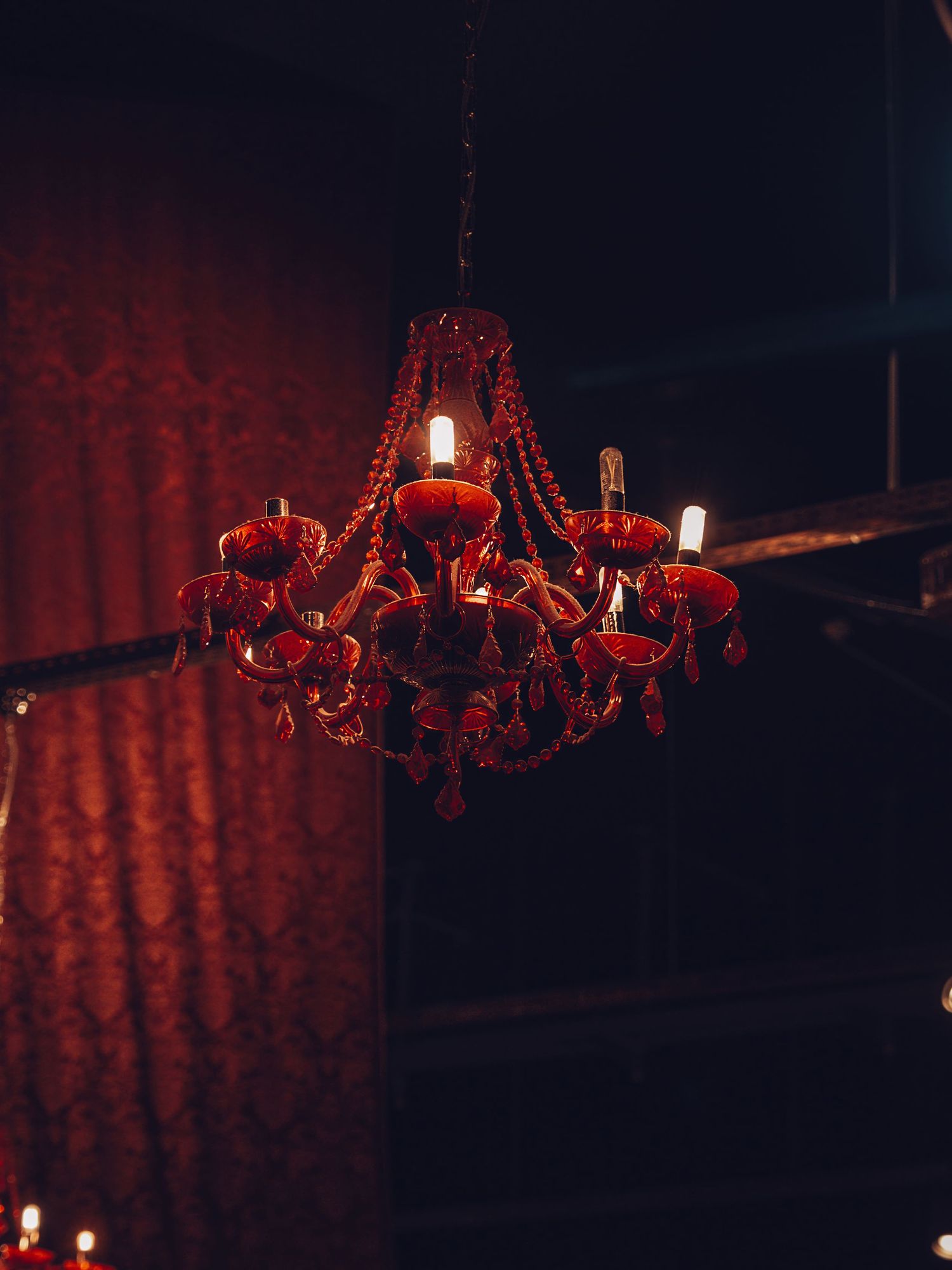A red crystal chandelier glows dimly in a dark room with dramatic lighting and red walls.