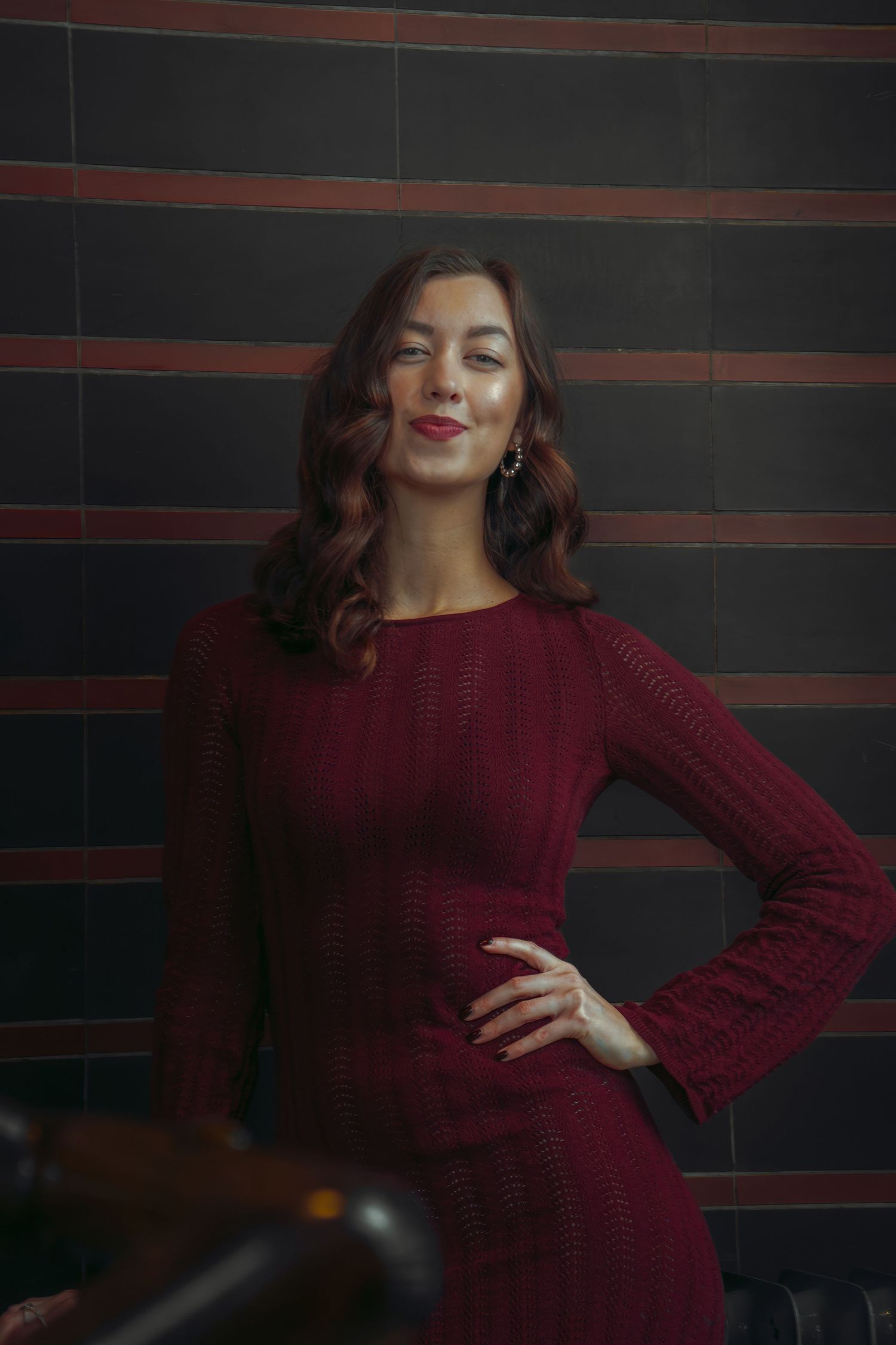 A woman in a burgundy knit dress poses against a dark background with horizontal lines.