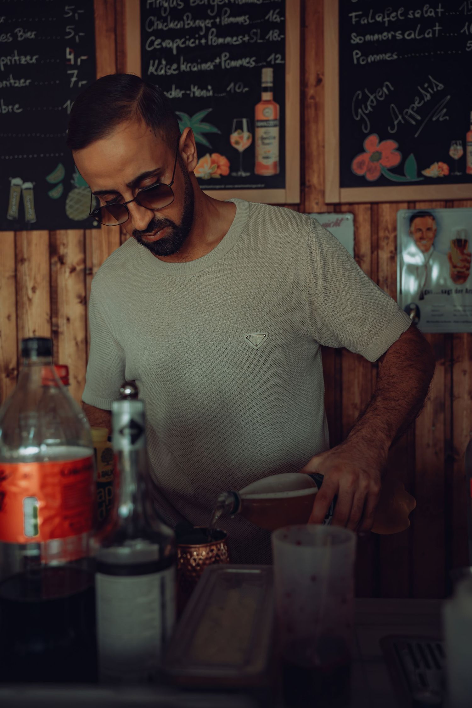 Bartender working behind wooden bar counter with chalkboard menu and bottles.