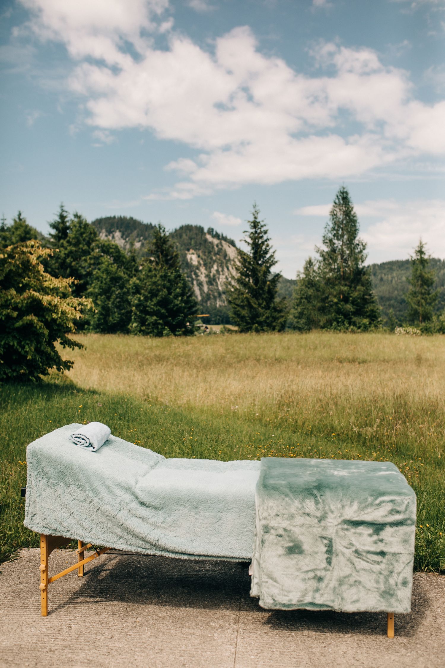 A massage table with gray cover sits outdoors against a scenic mountain landscape with evergreen trees and cloudy sky.