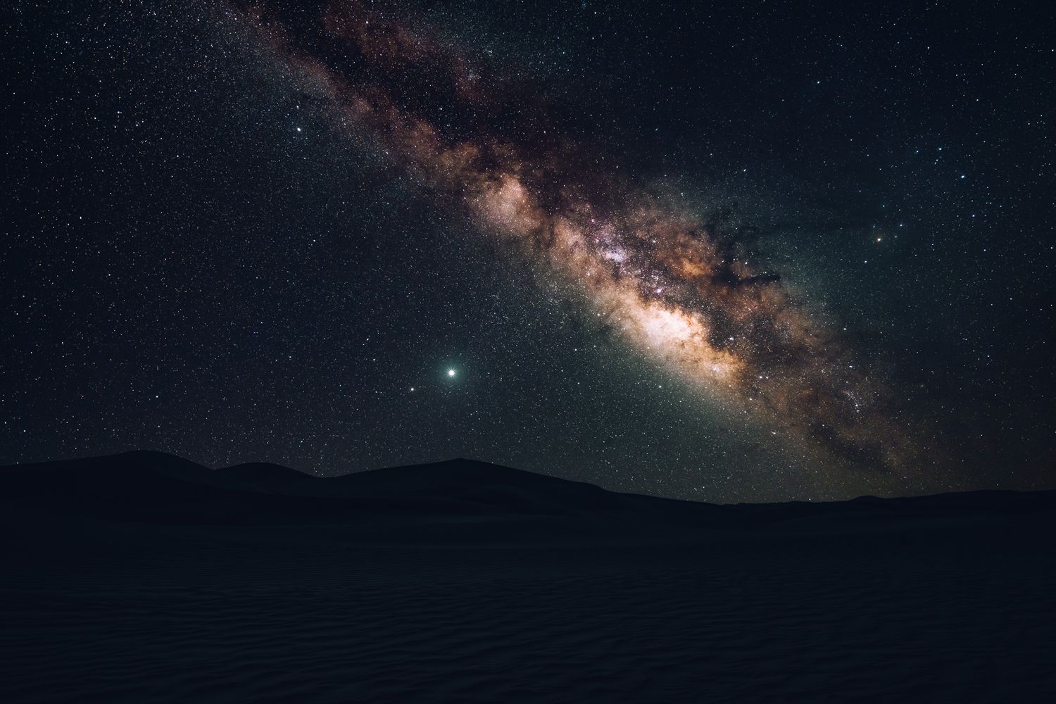 The Milky Way galaxy stretches across a dark night sky over mountain silhouettes in a stunning astrophotography scene.