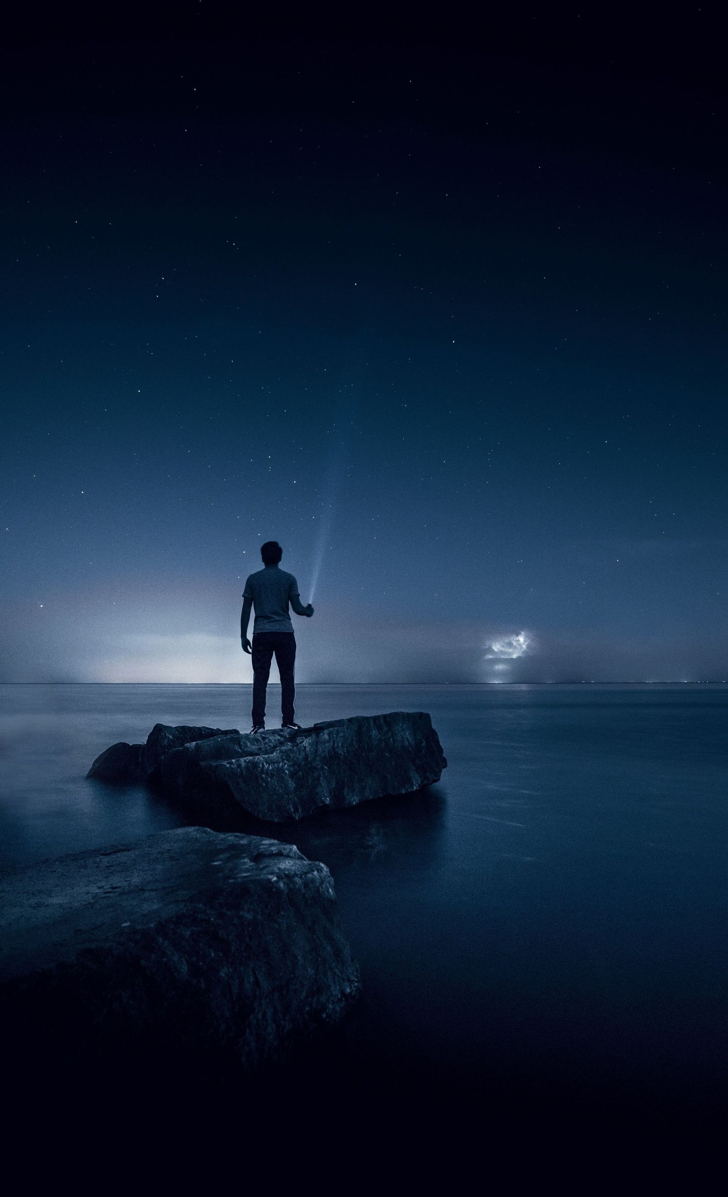 A silhouetted figure stands on a rock formation by the ocean at night holding a light beam against a starry sky.