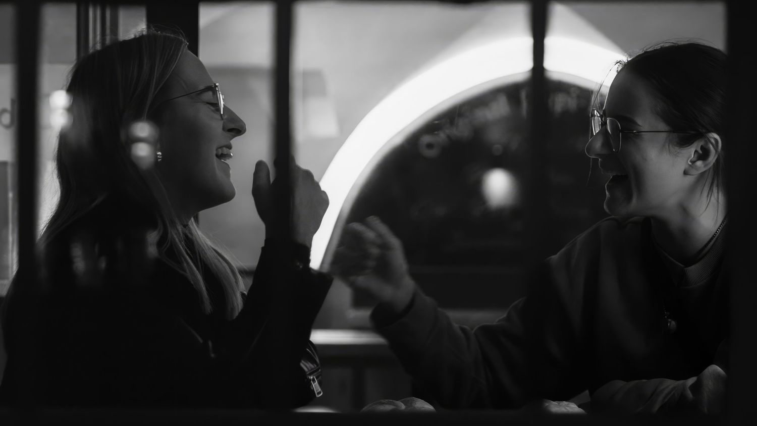 Two people sharing a candid laugh across a table in dramatic black and white lighting.