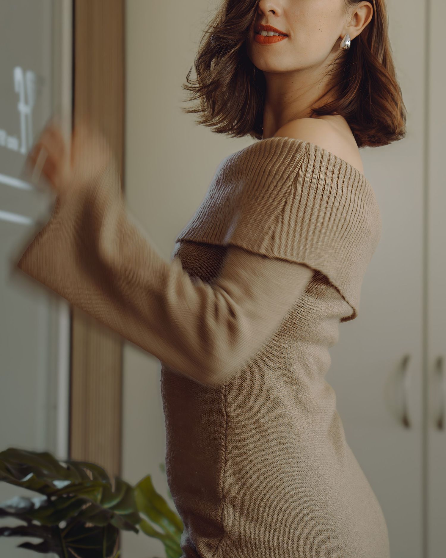 Elegant off-shoulder beige ribbed sweater photographed in soft natural lighting with houseplant in background.