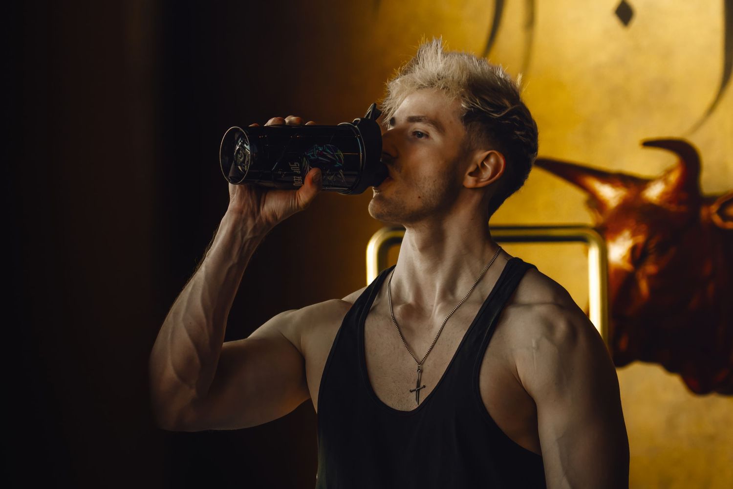 A person in a black tank top takes a drink from a dark bottle with warm lighting in the background. John Reed Linz. Manuell Stierberger
