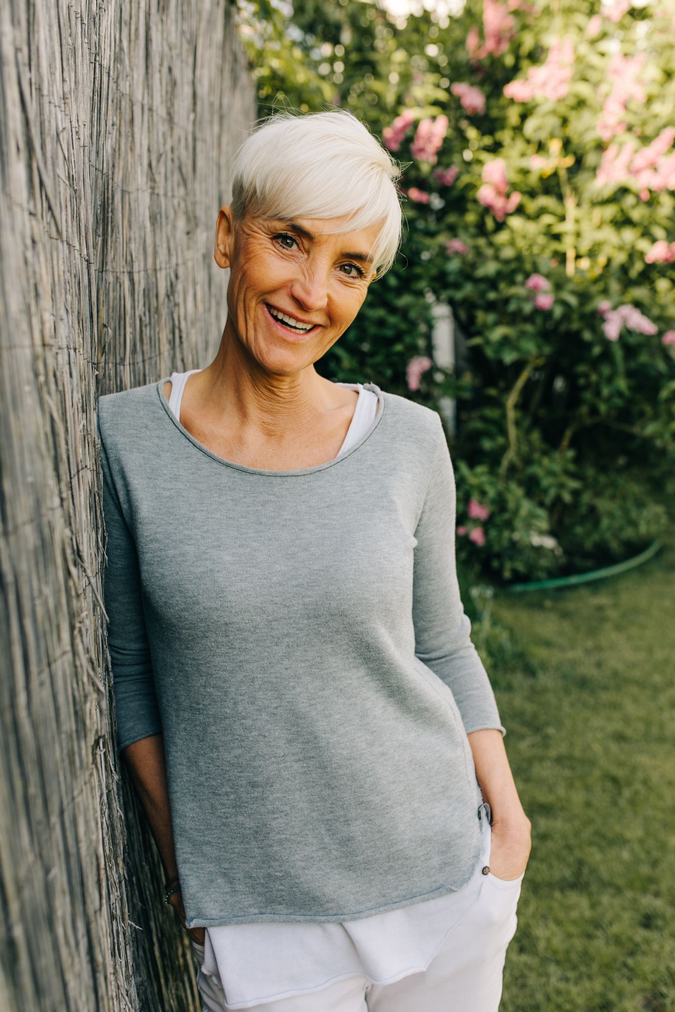 A person in a gray top and white pants stands by a wooden post in a garden setting.