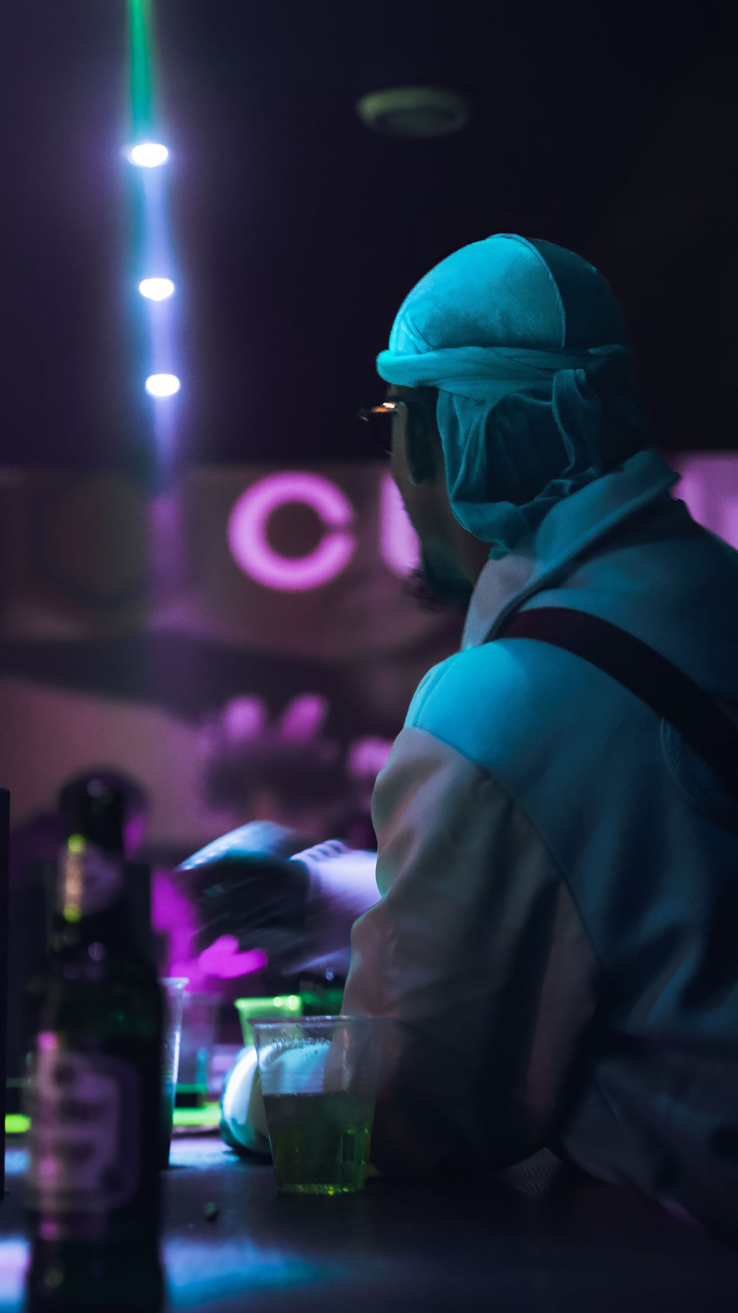 Person in baseball cap at bar counter with teal and purple ambient lighting.