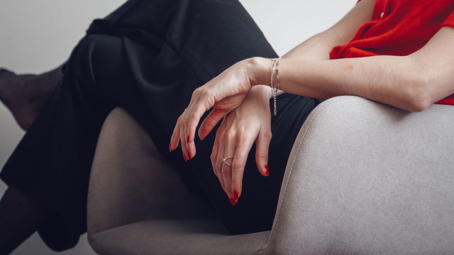 Close-up of hands resting on a gray couch with red nail polish against a neutral background.