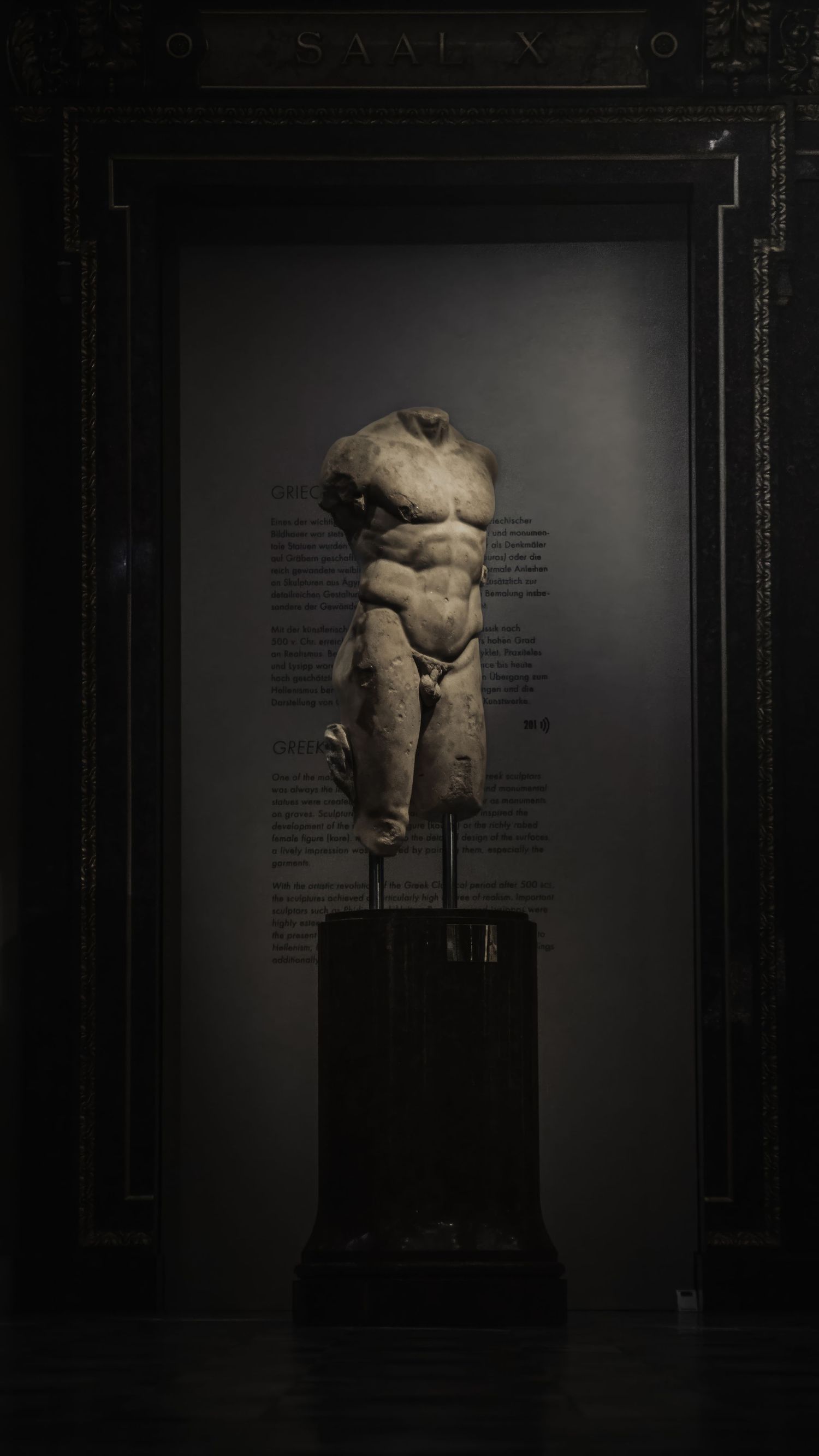 Ancient torso sculpture displayed in dramatic museum lighting against dark background.