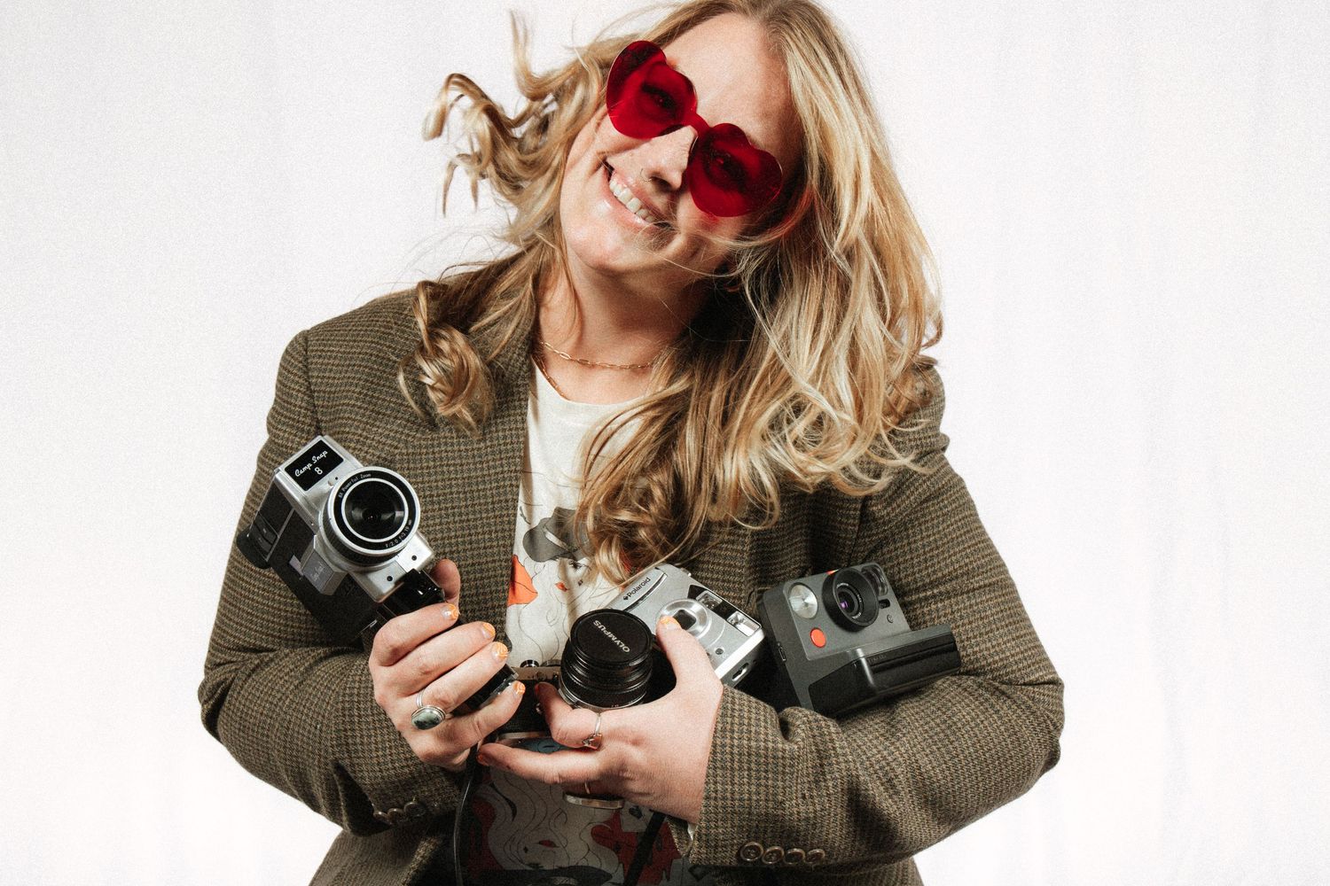 A photographer in a brown jacket holds multiple vintage cameras while wearing red sunglasses against a white background.