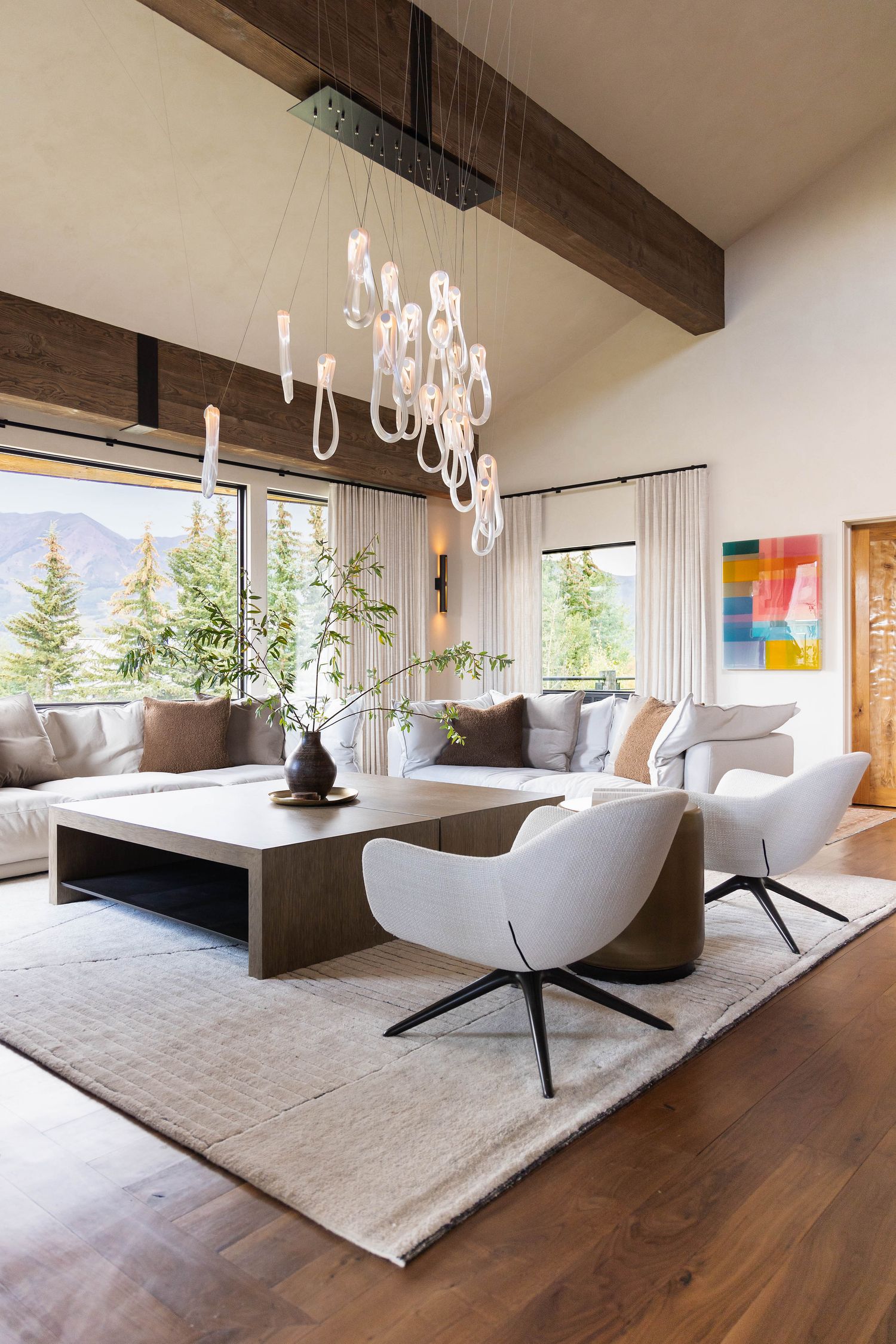Modern living room with white chairs, dark wood coffee table, statement chandelier, exposed beams, and mountain views through large windows.