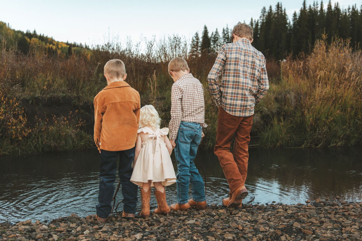 Four children stand at a rocky riverbank surrounded by autumn foliage, viewed from behind in casual fall attire.