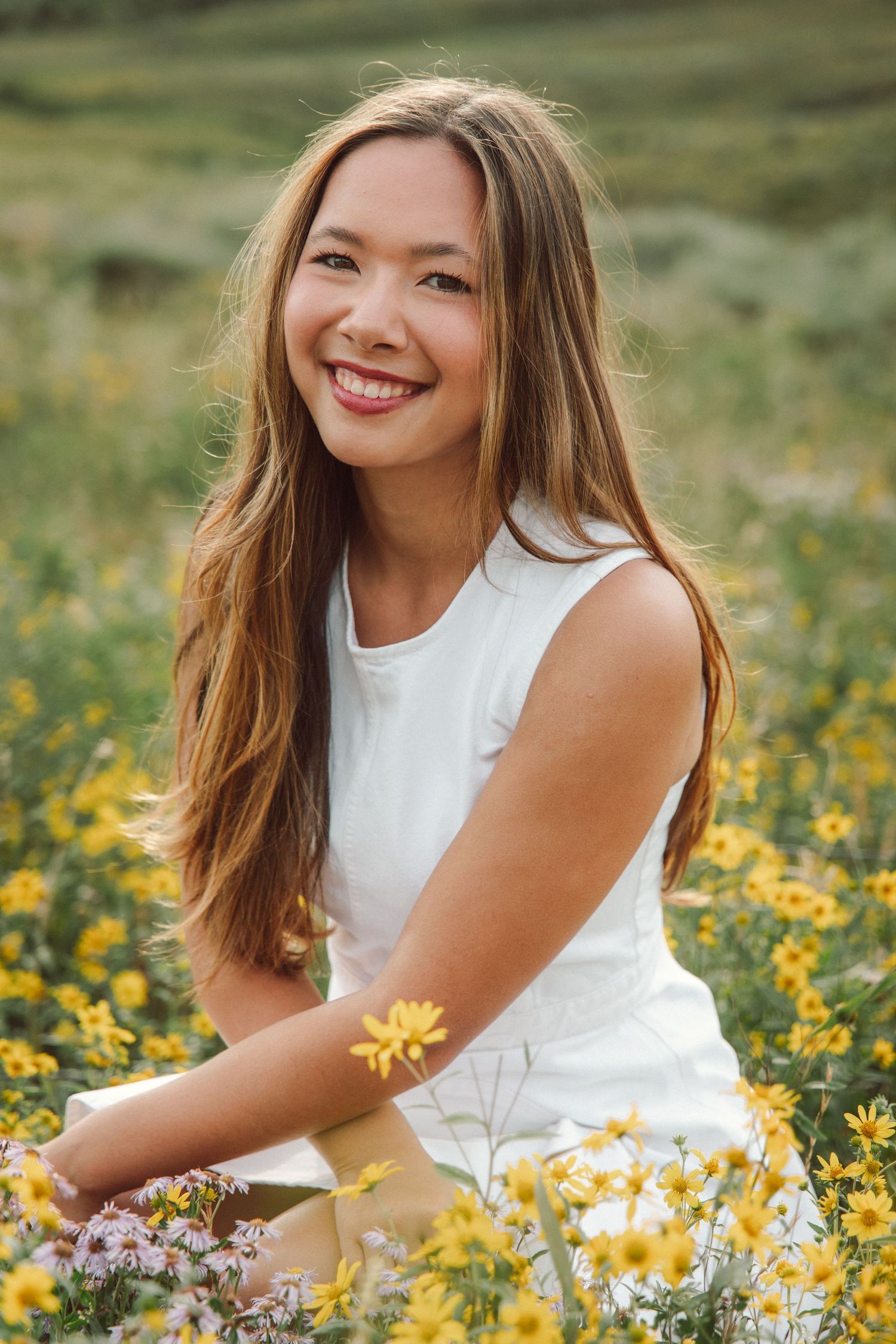 Smiling young woman with long hair sitting in a field of yellow wildflowers wearing a white sleeveless top.