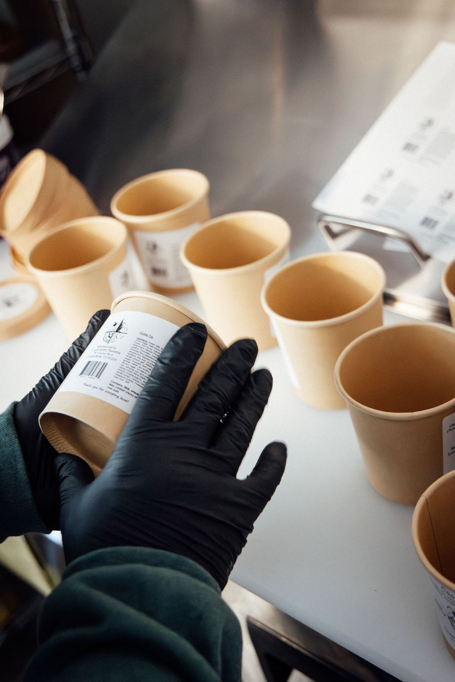 Hands in black gloves labeling kraft paper cups on a white surface in a food service setting.