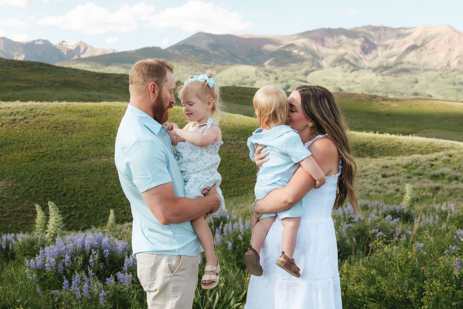Family of four in matching blue and white outfits posing in a wildflower meadow with mountain views.