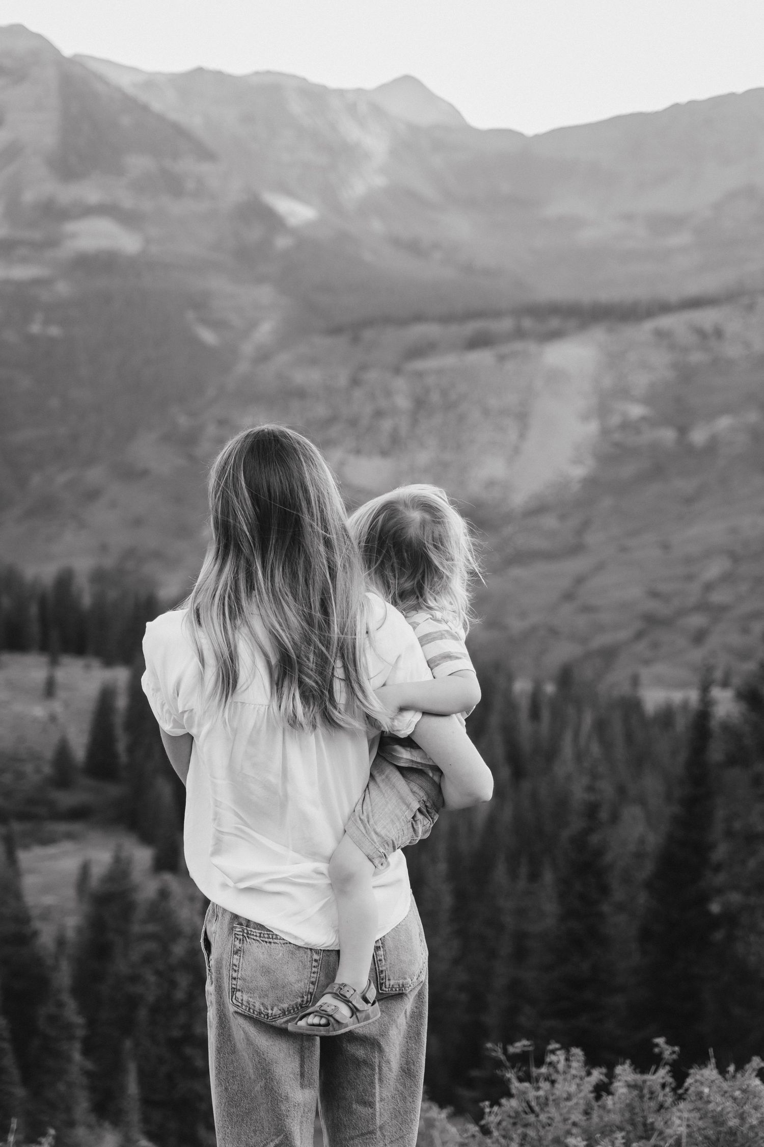 Black and white photo of a mother holding her child, gazing at a stunning mountain landscape with pine trees.