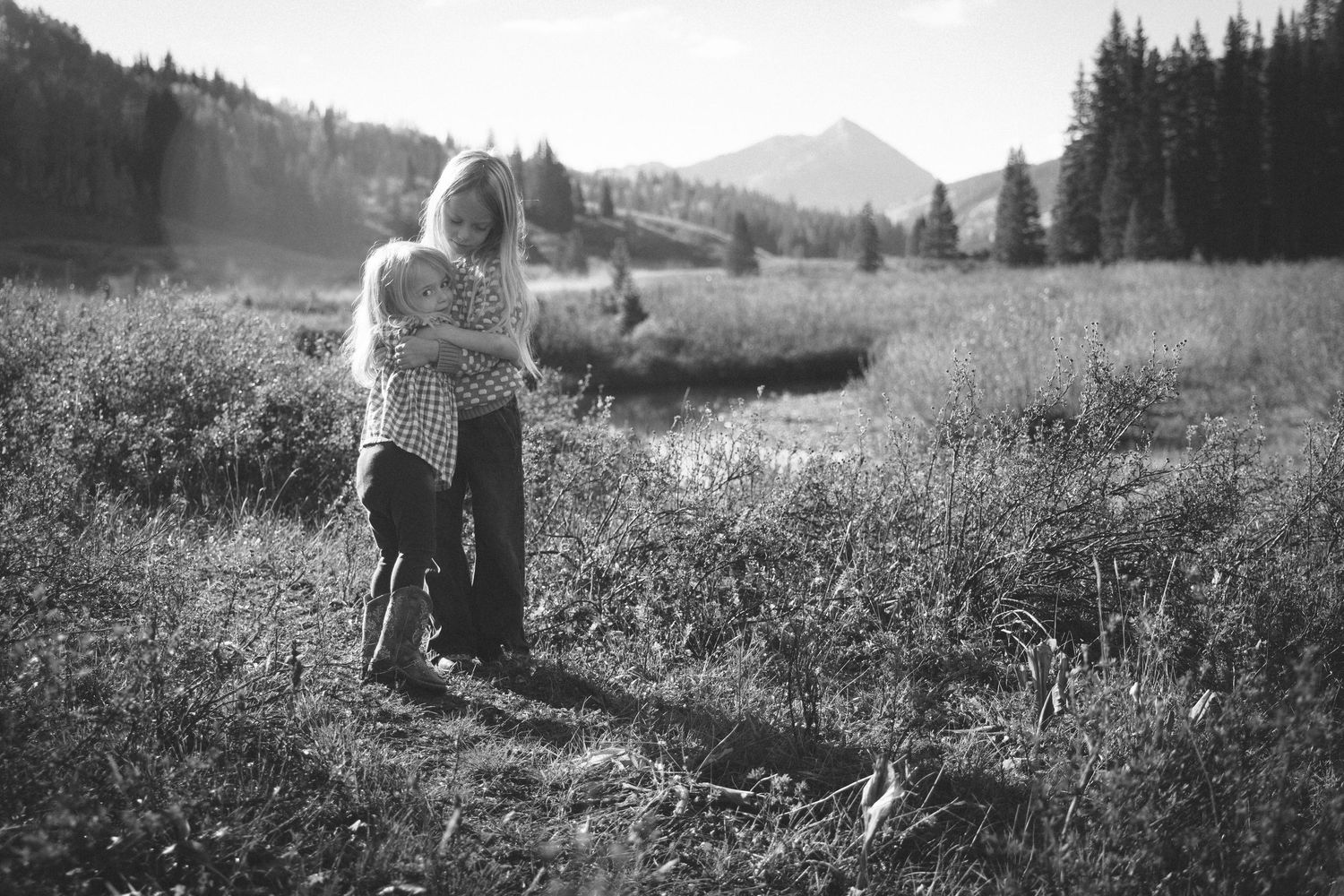 Black and white photo of a mother and child embracing in a meadow with mountains and pine trees in the background.