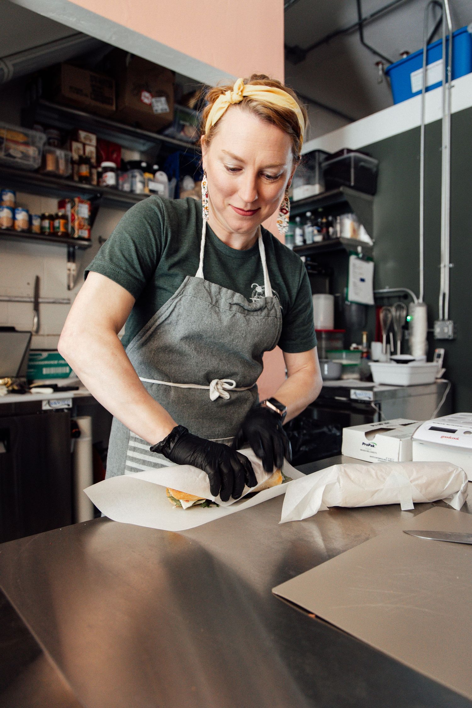 Female chef in gray apron and black gloves wrapping a sandwich in parchment paper in a commercial kitchen.