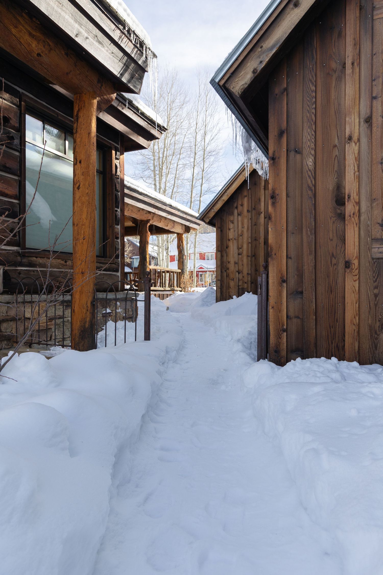 Snow-covered pathway between rustic wooden cabin buildings with bare aspen trees visible against a winter sky.