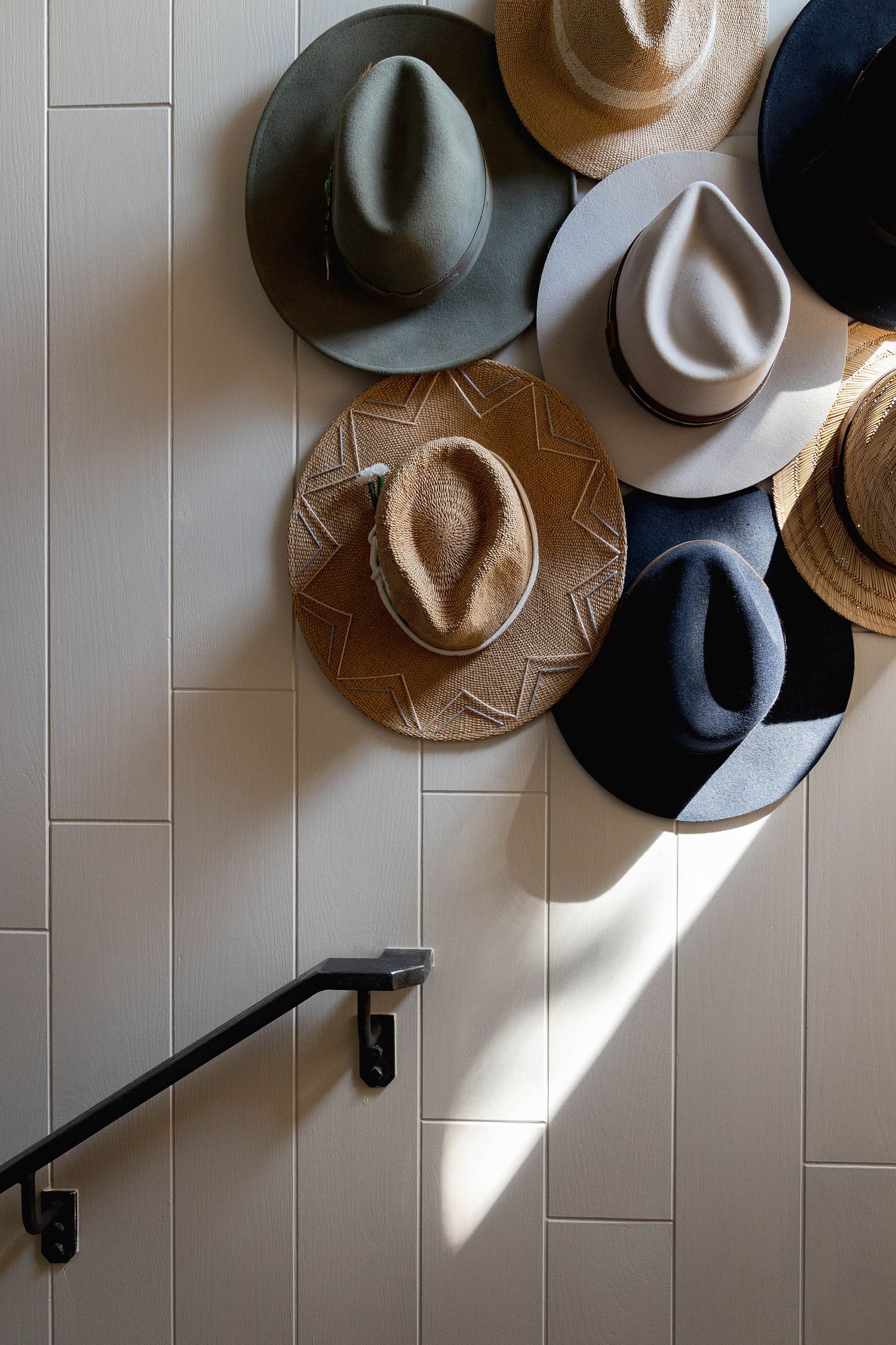 Collection of stylish fedora hats in neutral tones displayed on a white tiled wall above a black staircase railing.