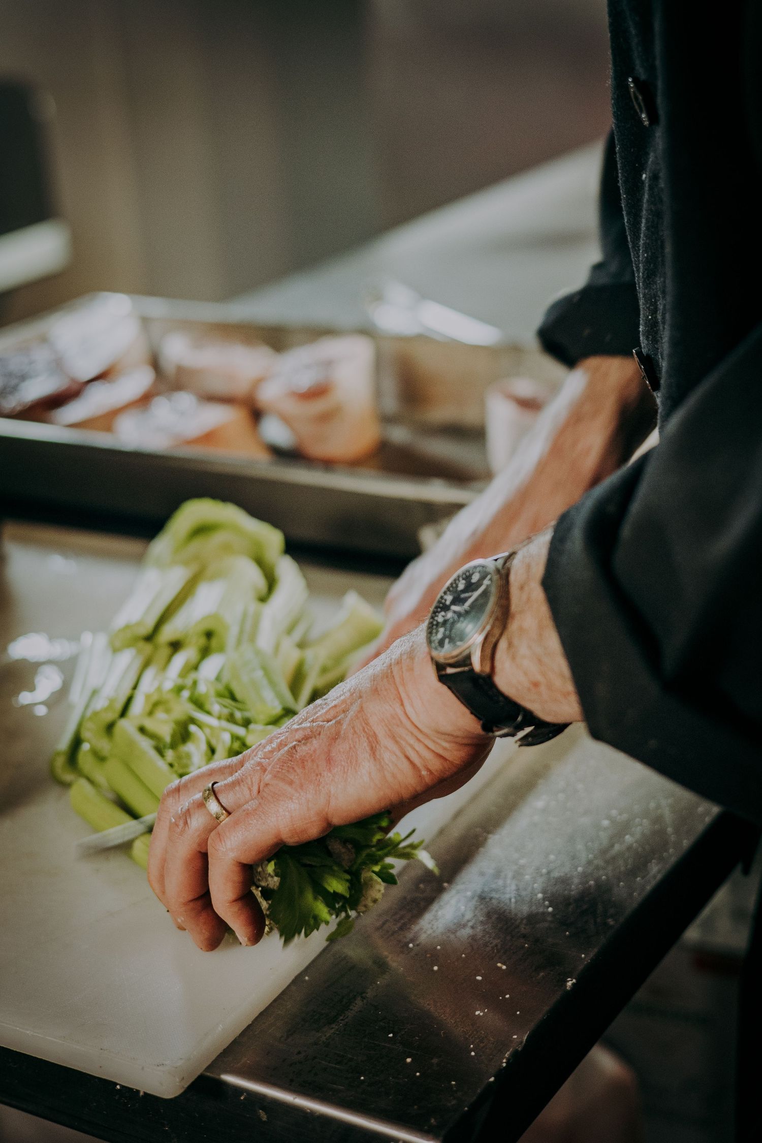 A chef wearing a black sleeve and watch chops fresh celery on a white cutting board in a professional kitchen.