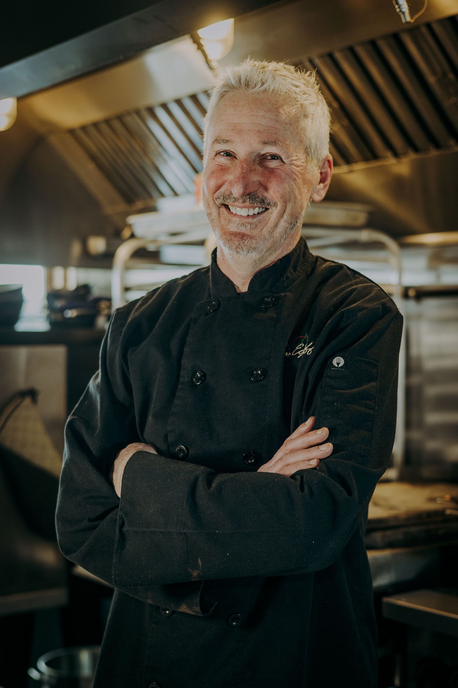 Professional chef in black uniform smiling in commercial kitchen setting.