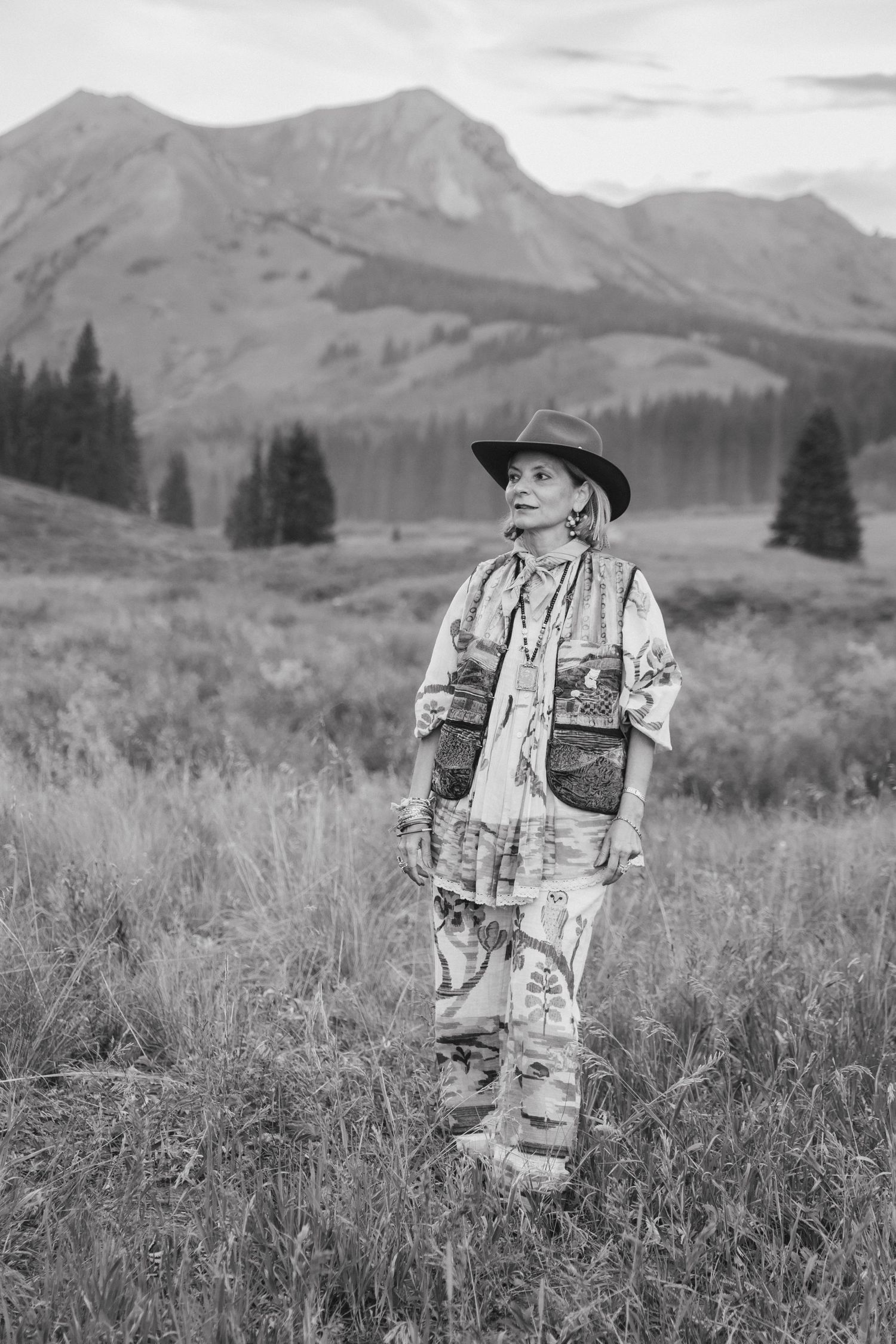 Black and white portrait of a woman in a wide-brim hat and embellished vest standing in a grassy mountain meadow with evergreen trees.