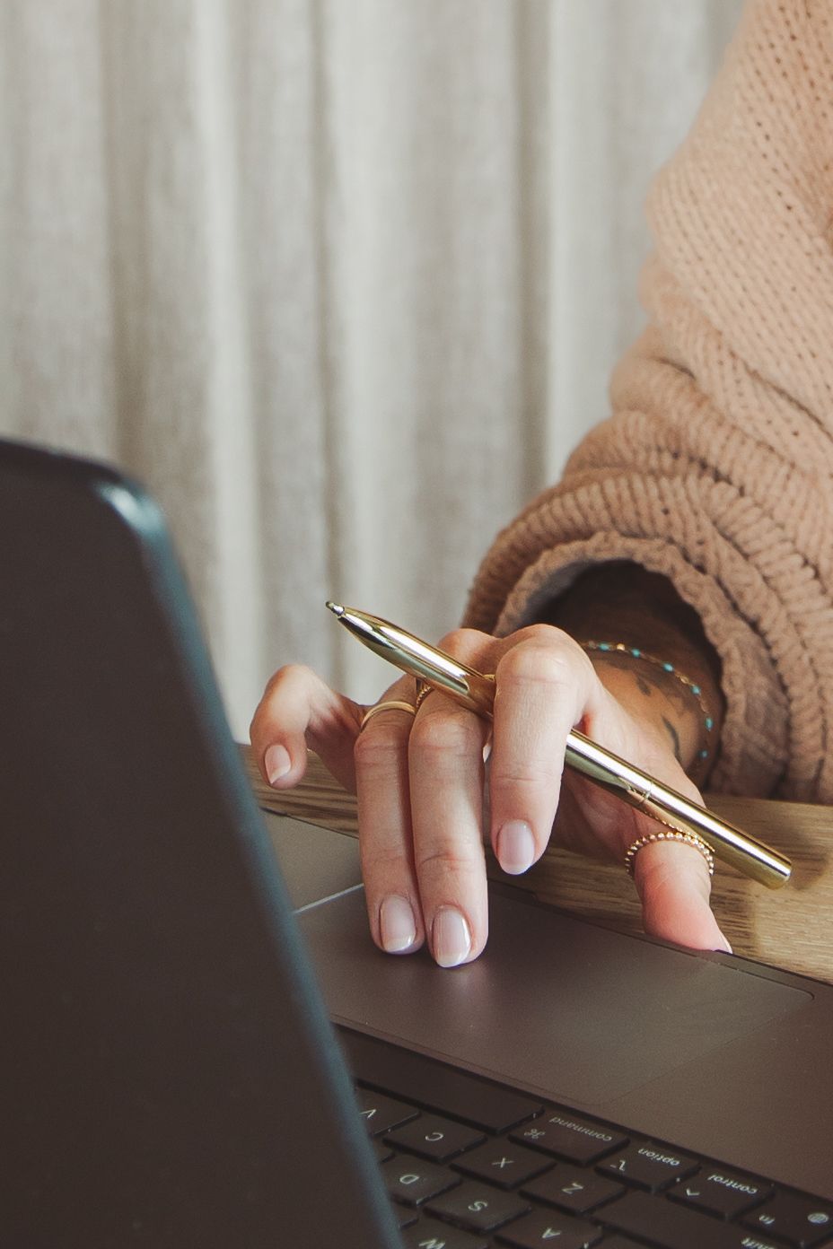 Woman in beige knit sweater holding a gold pen over a laptop keyboard with a notebook nearby.