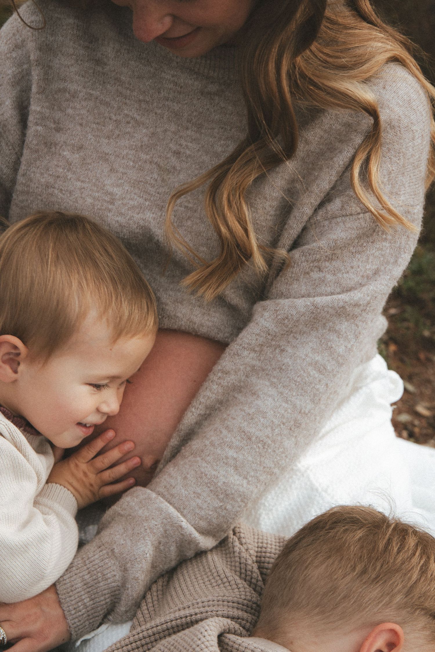 Pregnant woman in beige sweater sitting outdoors with two young toddlers leaning against her baby bump.