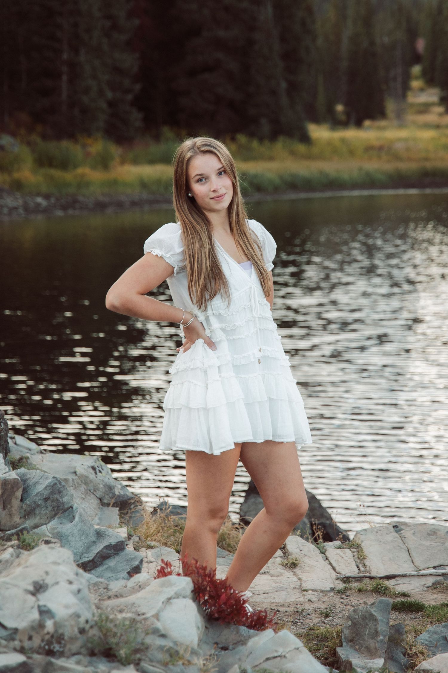 Person in white dress poses on rocky shoreline near a serene lake.