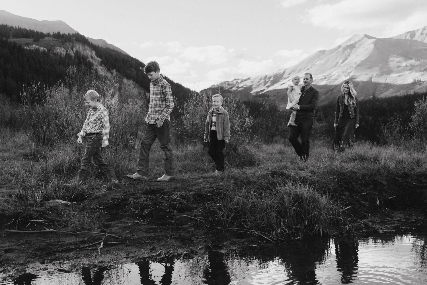 Black and white photo of five people standing on a grassy hillside with mountain peaks and a reflective pond in background.