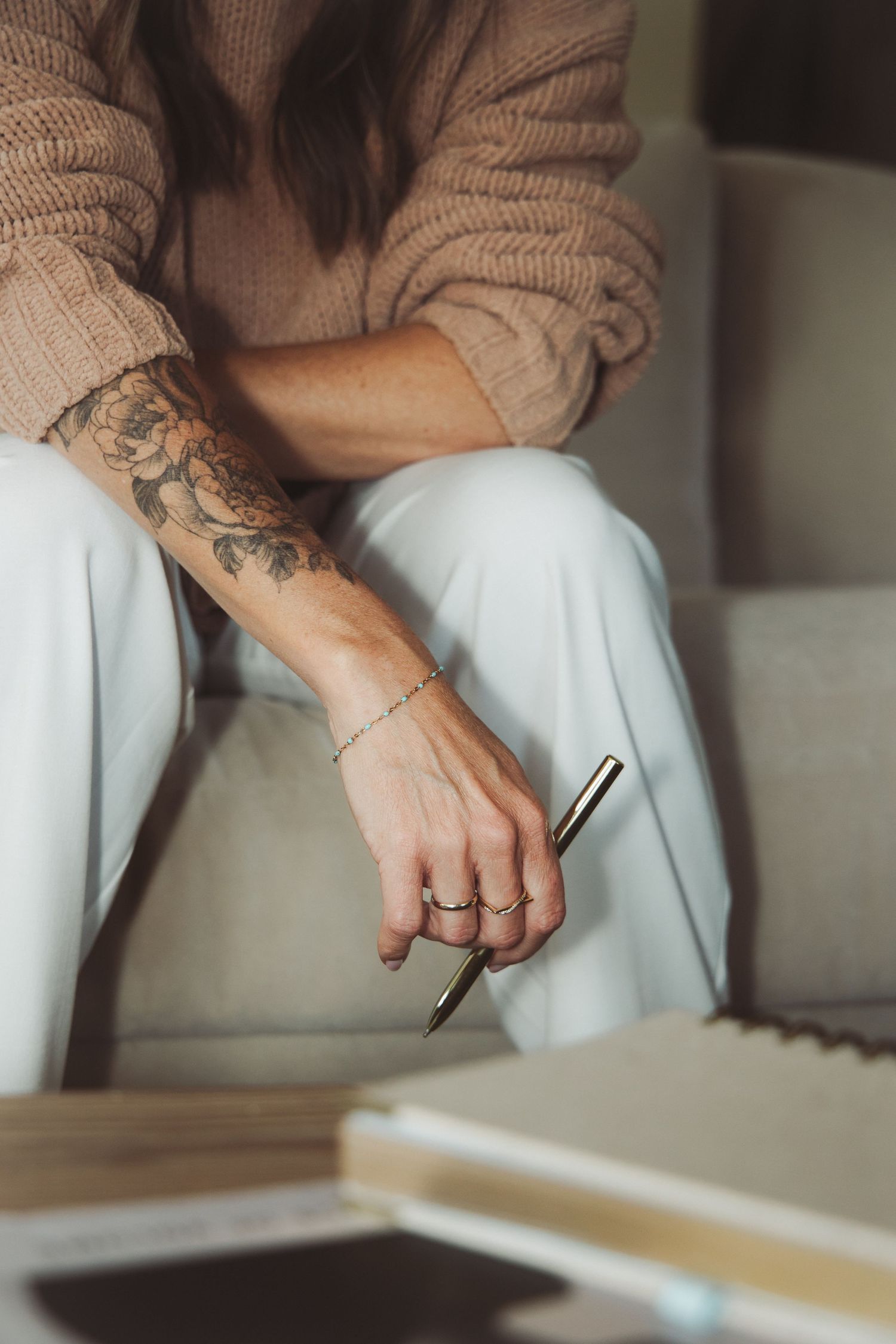 Woman with floral tattoo and rings holds a gold pen while seated on a light sofa wearing white pants.