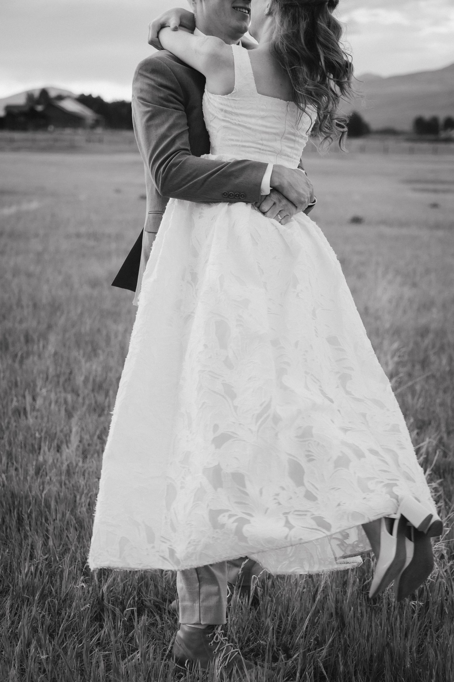 Black and white photo of bride and groom embracing in a field, bride wearing tea-length lace wedding dress.