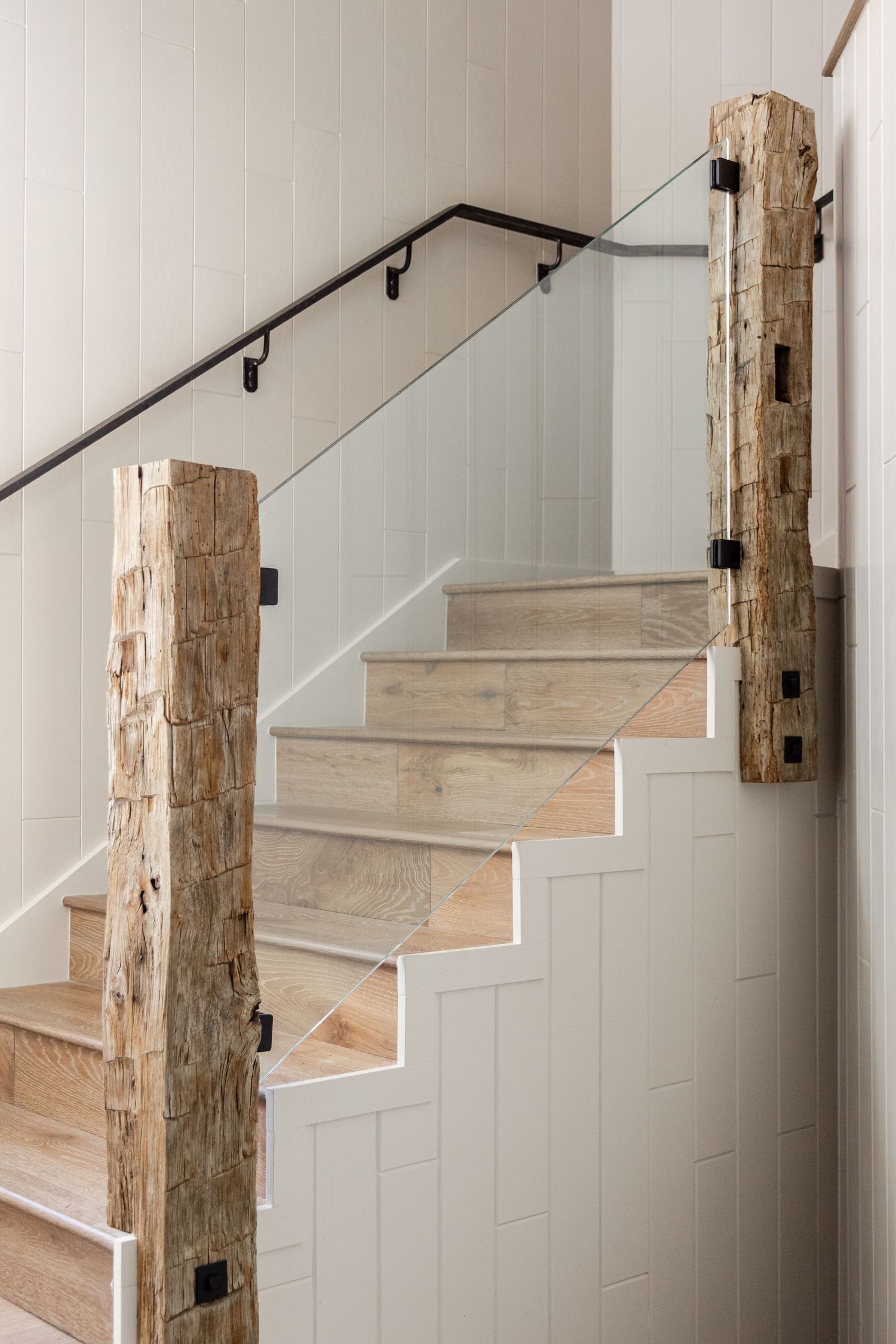 Modern staircase with rustic wooden log newel posts, glass panel railing, light wood steps, and white shiplap walls.