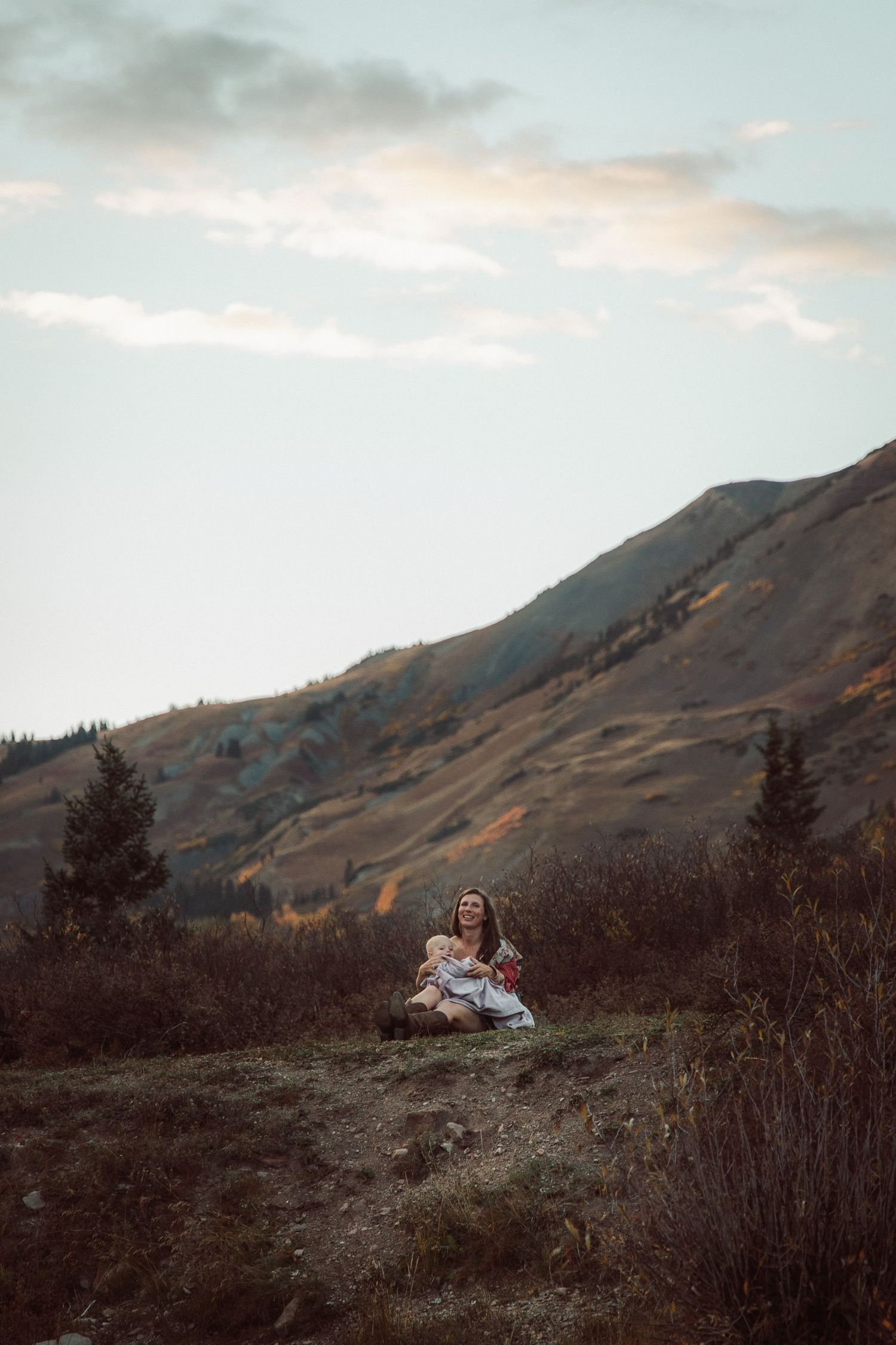 Two people sit together on a rocky mountain overlook at sunset with dramatic mountain ranges in the distance.