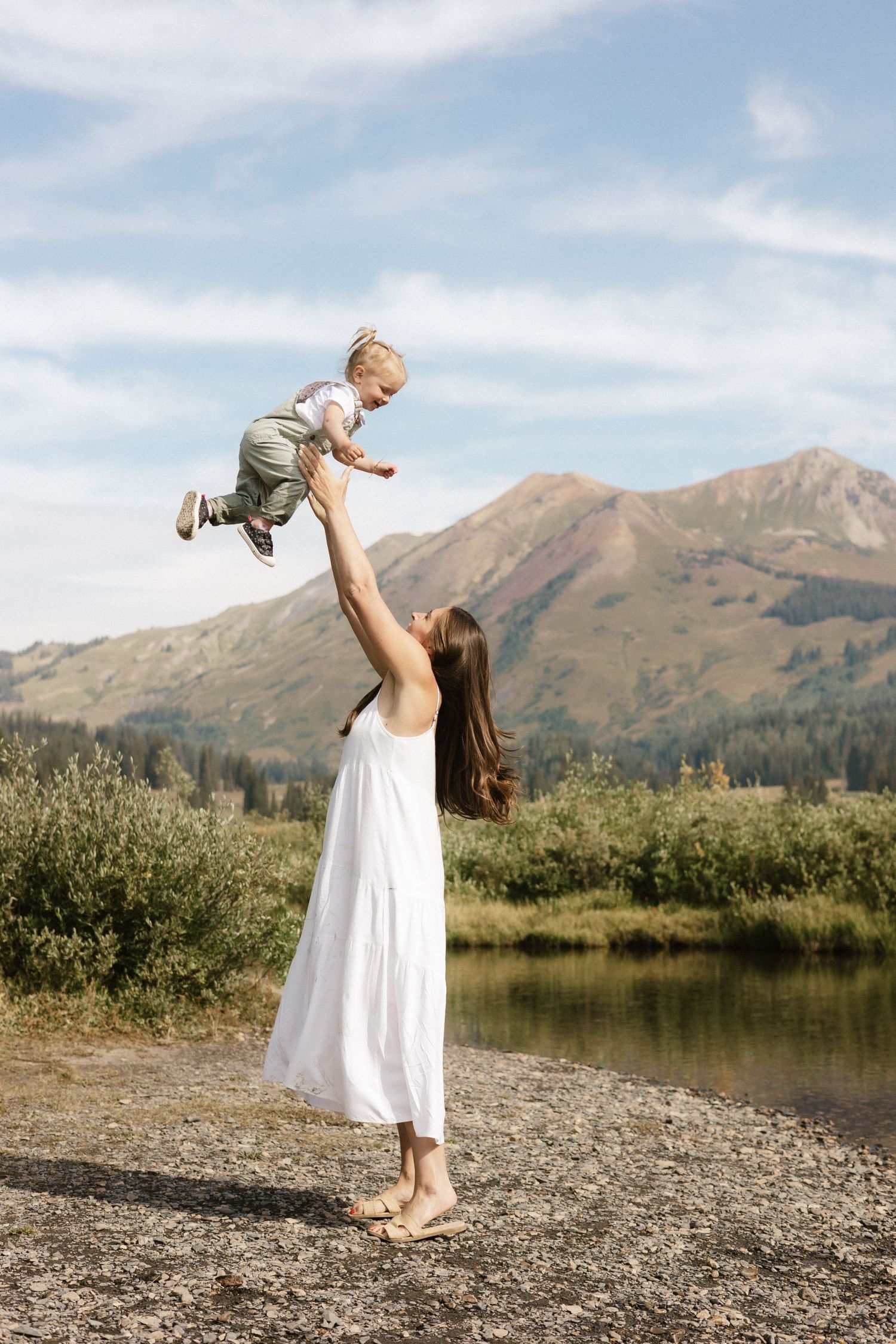 A magical moment by a mountain lake as a white-dressed figure lifts a child against the scenic backdrop.