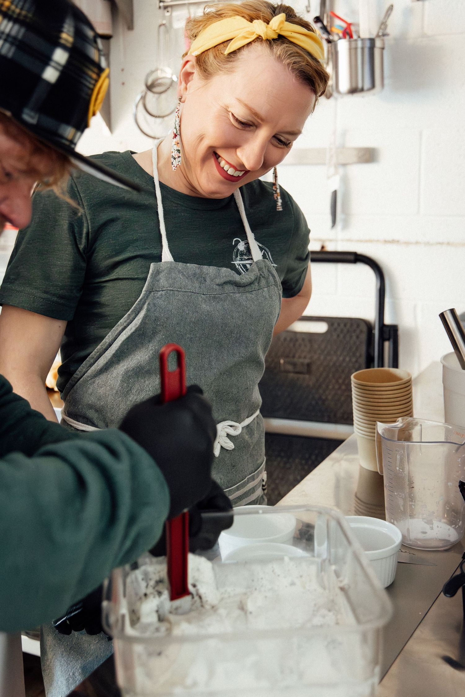 Smiling woman in gray apron and yellow headband working with ice in a commercial kitchen.