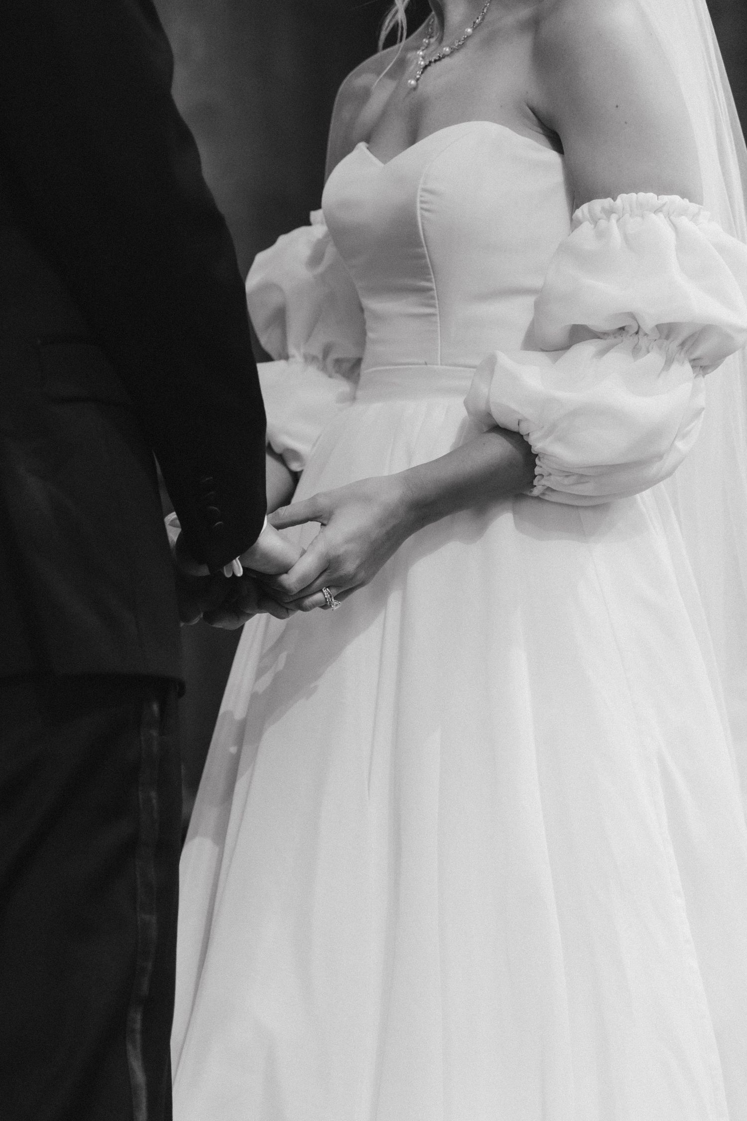 Black and white photo of bride in strapless gown with puff sleeves holding hands with groom at wedding ceremony.