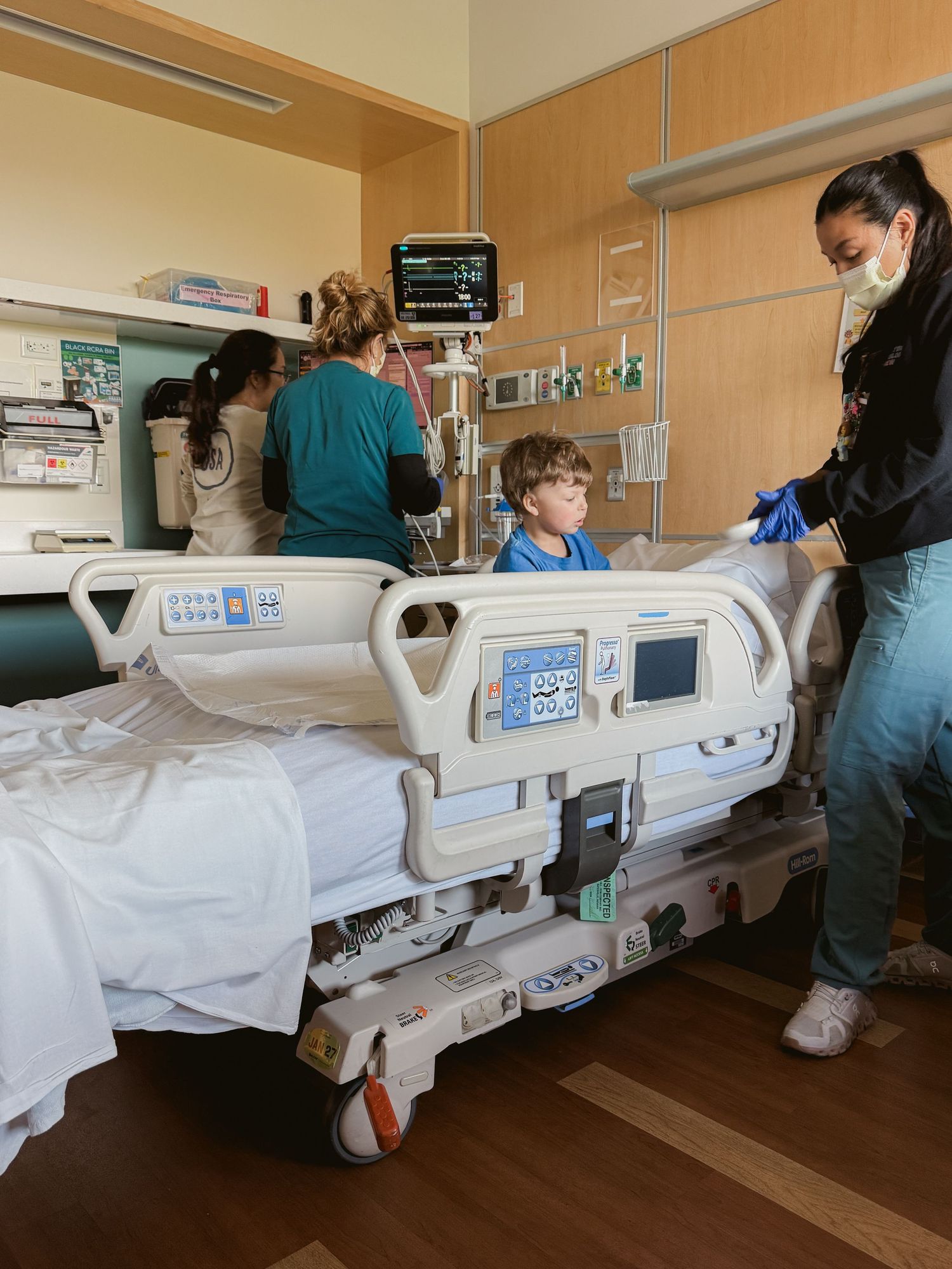 Medical staff and family assist a young boy in a hospital room with an adjustable bed and monitoring equipment.