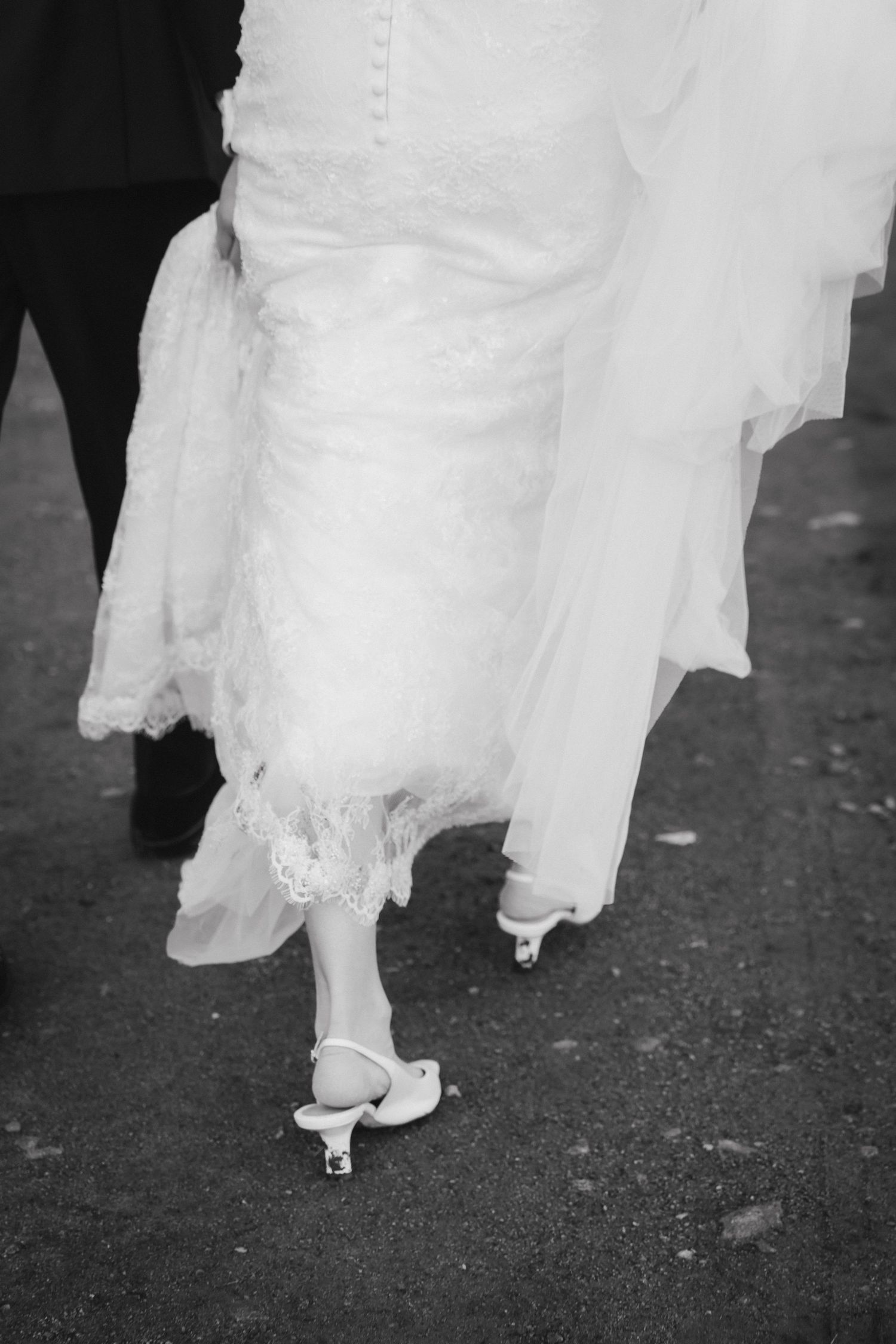 Black and white photo of a bride in a lace wedding dress and white heels walking on pavement beside a groom in dark trousers.