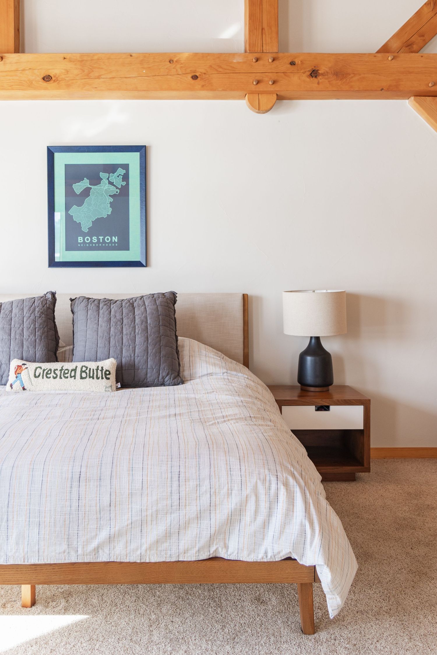Cozy bedroom with wooden beams, striped bedding, gray pillows, a Boston map print, and a modern nightstand with lamp.